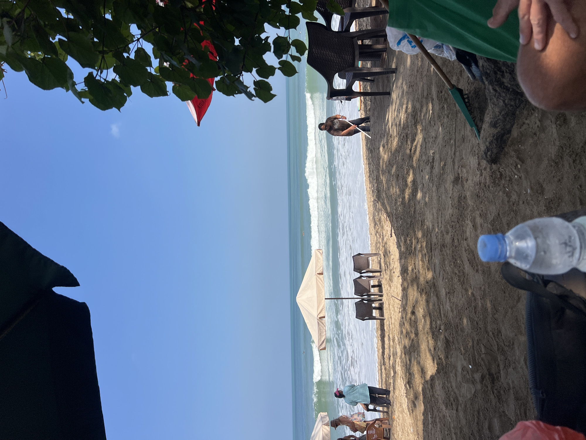 The image depicts a beach scene in Kuta, Indonesia, likely on a sunny day with clear blue skies. In the foreground, a dark umbrella casts a shadow over the sandy beach. A bottle of water with a blue cap is prominently placed. Further back, several beach chairs are arranged under a light-colored umbrella. The ocean is visible with gentle waves, and a few surfers can be spotted in the distance. A person, dressed in a dark t-shirt and pants, is seen raking the sand near the water's edge. Another person, wearing a blue top and a pink hat, stands by the beach chairs, facing the ocean. There are other individuals in the background, some appearing to be beachgoers and others possibly working on the beach. The overall atmosphere suggests a typical day at a popular tourist destination.