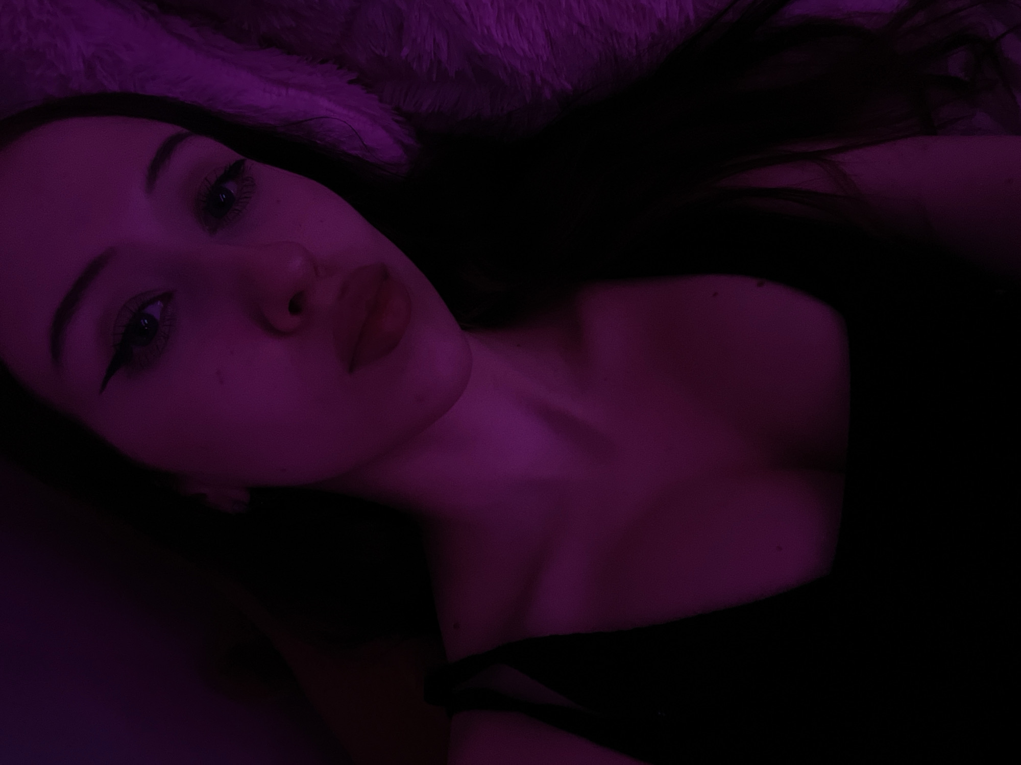 A young woman with dark hair and winged eyeliner is shown in a close-up selfie. She is wearing a black tank top and is illuminated by purple light. The background is dark and appears to be a soft, textured surface. The location is the city of Votkinsk, Russia.