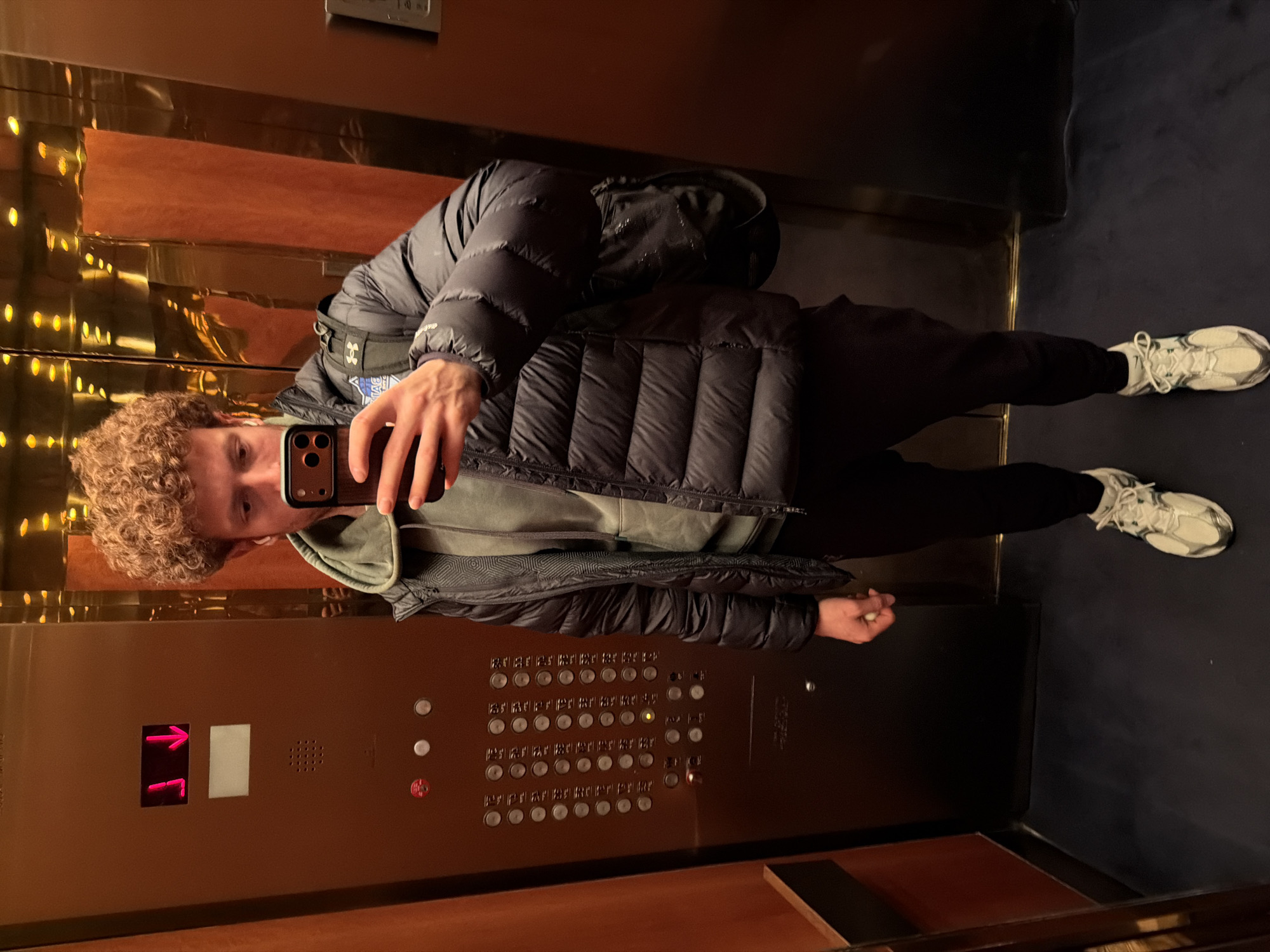 The image shows a man taking a selfie in an elevator. He is wearing a black puffer jacket, a green hoodie, black pants, and white sneakers. He has curly hair. The elevator has brown walls and a panel with buttons for multiple floors. The floor of the elevator is blue. Based on the extensive floor selection panel, it is likely in New York City, United States of America.