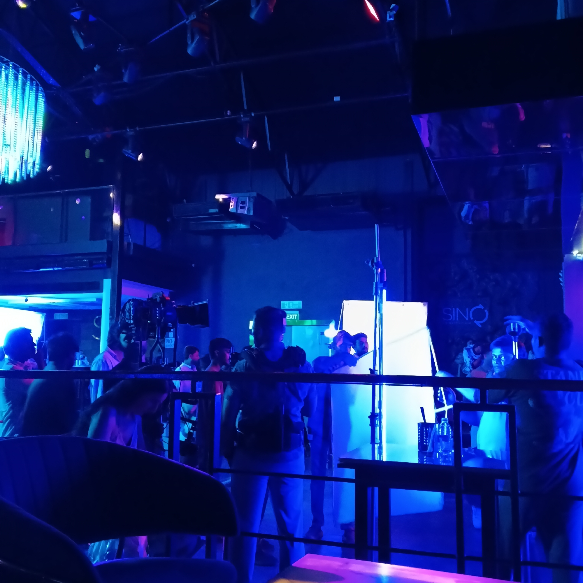 The image shows an interior scene at a bar in Sinquerim, India. The bar appears to be a nightclub or lounge, with a blue light atmosphere, possibly from LED fixtures or lighting effects. A bar area is visible in the foreground and an upper level with a reflective surface.
Several people are present, but they are mostly obscured or in silhouette. Some are standing and conversing, while others appear to be involved in setting up lighting equipment. There is a light panel or reflector supported by a stand.
The word "EXIT" is seen in the background. The name "SINO" is visible on the wall. There is a chandelier in the background.