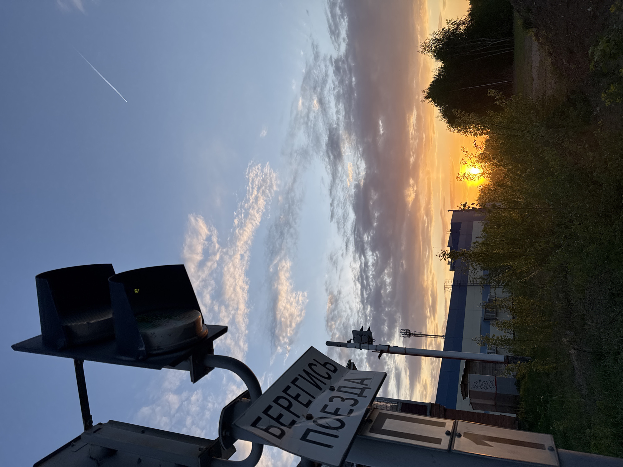 The image shows a railway crossing in the city of Chaikovsky, Russia at sunset. The sun is setting behind trees and a large industrial building. A contrail is visible in the upper left of the sky. In the foreground is a railroad crossing signal with a sign in Russian that reads "БЕРЕГИСЬ ПОЕЗДА" (Beware of the Train). The signal has two black hoods covering signal lights. A cellular tower is also visible near the building. The image is taken from a low angle, looking upwards towards the sky and setting sun.