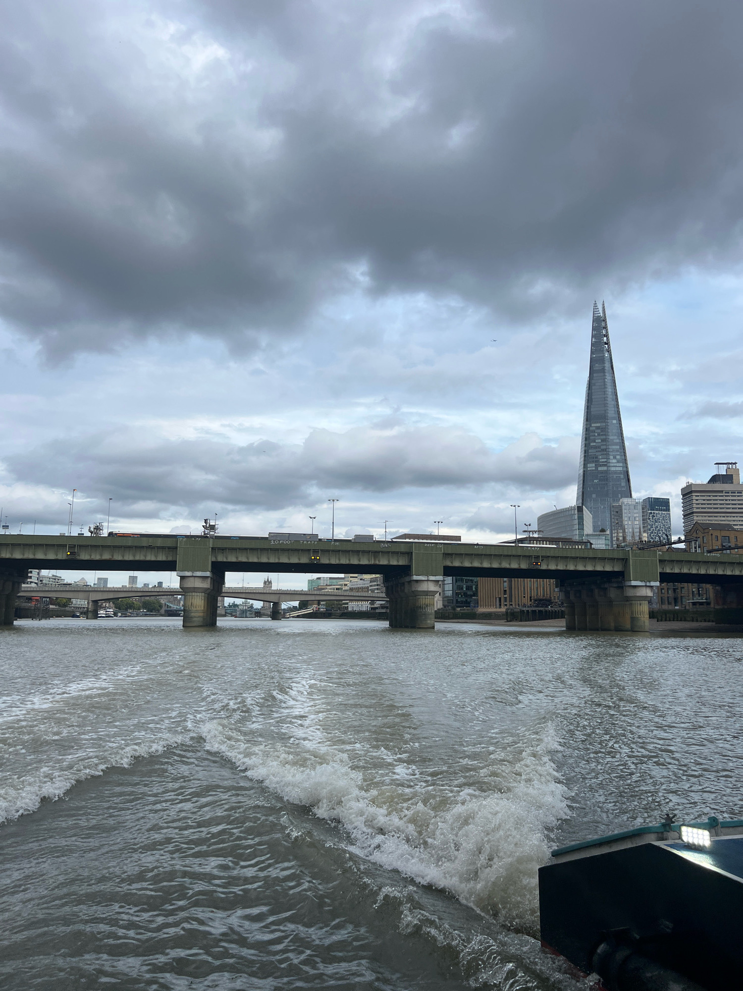 The image depicts a view from a moving boat on the River Thames in London, United Kingdom. In the foreground, white foamy waves churn behind the boat, indicating its forward motion. The river is a muted grey-brown color, reflecting the overcast sky.
Dominating the midground is a wide, multi-lane road bridge, with concrete pillars supporting its structure. Vehicles are visible on the bridge, including trucks. In the distance, beyond the bridge, the iconic modern skyscraper, The Shard, pierces the cloudy sky. Several other buildings of varying architectural styles are also visible along the London skyline. The sky is filled with heavy, grey clouds, suggesting a cool or potentially wet day. The overall impression is of a utilitarian, urban landscape viewed from the water.