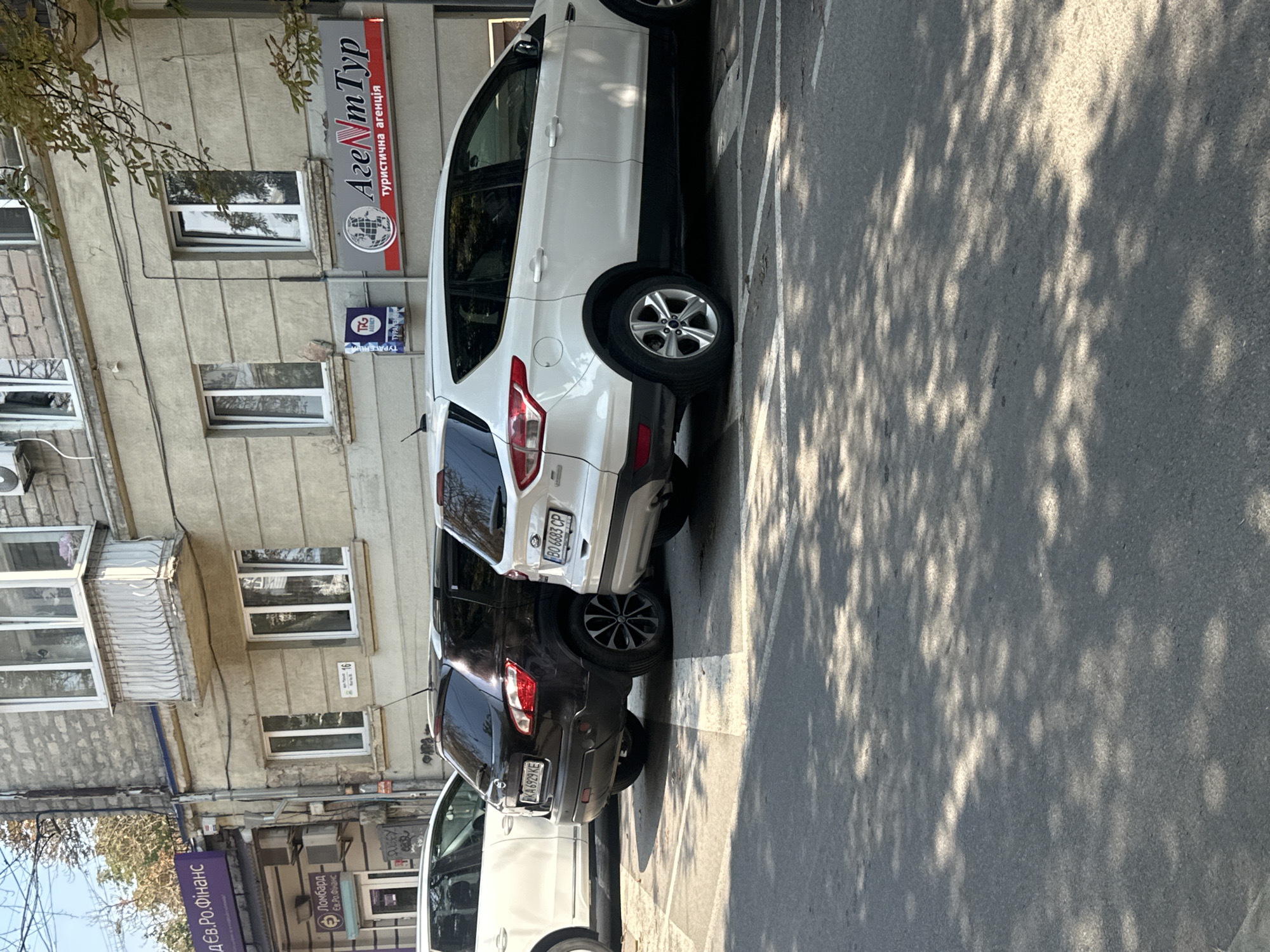 The image shows a street scene in Ternopil, Ukraine, with several cars parked along a building. The building appears to be a commercial establishment, with signs visible for "ДЕВ.РО.ФІНАНС" (DEV.RO.FINANCE), "Ломбард" (Pawnshop), and "АгентТур" (AgentTour) a travel agency. The cars parked are a white SUV, a dark colored SUV, and a white sedan. The street is paved with asphalt and dappled with sunlight and shadows from trees. The building is light beige with several windows. License plates are visible on two of the cars. The overall impression is a typical urban street scene.