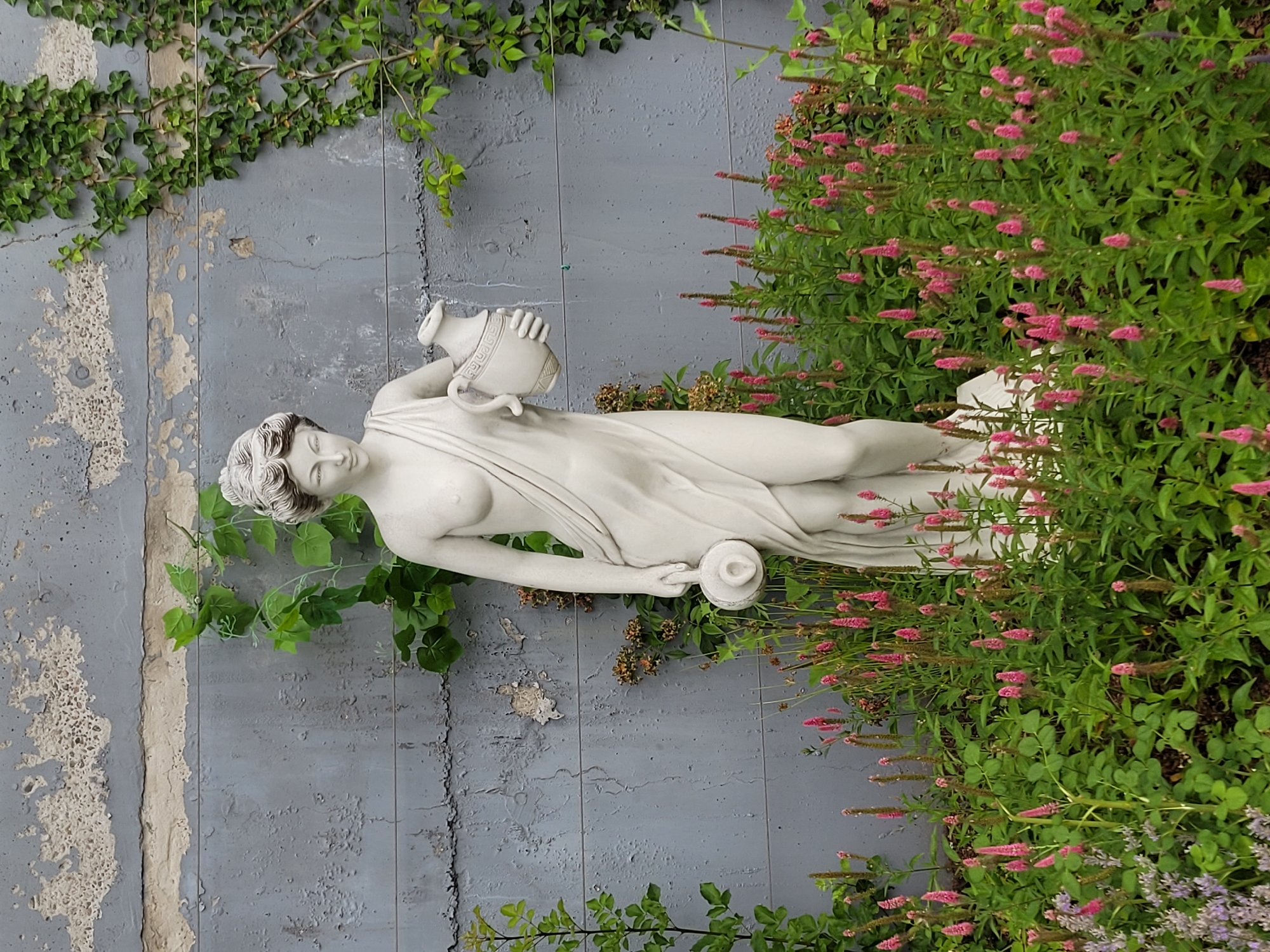 A white marble statue of a woman stands in a garden in front of a grey, textured concrete wall. The statue depicts a female figure wearing a draped garment, with her hair styled in an updo. She is holding a jug in her left hand and a smaller vessel in her right hand. The garden is filled with lush green foliage and numerous pink flowers. Green ivy vines grow on the concrete wall behind the statue. The scene is set outdoors, with natural lighting.