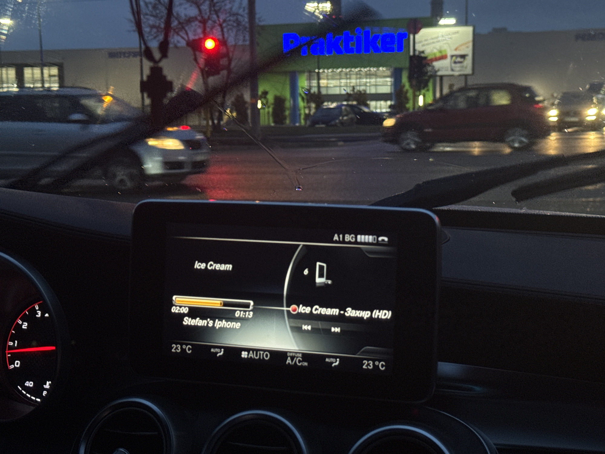 The image, taken from inside a car, shows a nighttime scene. The car's dashboard is visible, displaying a screen with music playing, titled "Ice Cream - Захир (HD)" from "Stefan's Iphone". The temperature is set to 23°C. Outside the car, through the rain-streaked windshield and activated wipers, a busy street scene unfolds. A red traffic light is lit. The "Praktiker" store is visible in the background. Cars are driving on the road. Based on the "A1 BG" indicator on the dashboard screen, the location is likely in Bulgaria. Given the Praktiker store and the Cyrillic alphabet in the song title, it's further pinpointed to the city of Sofia, Bulgaria.