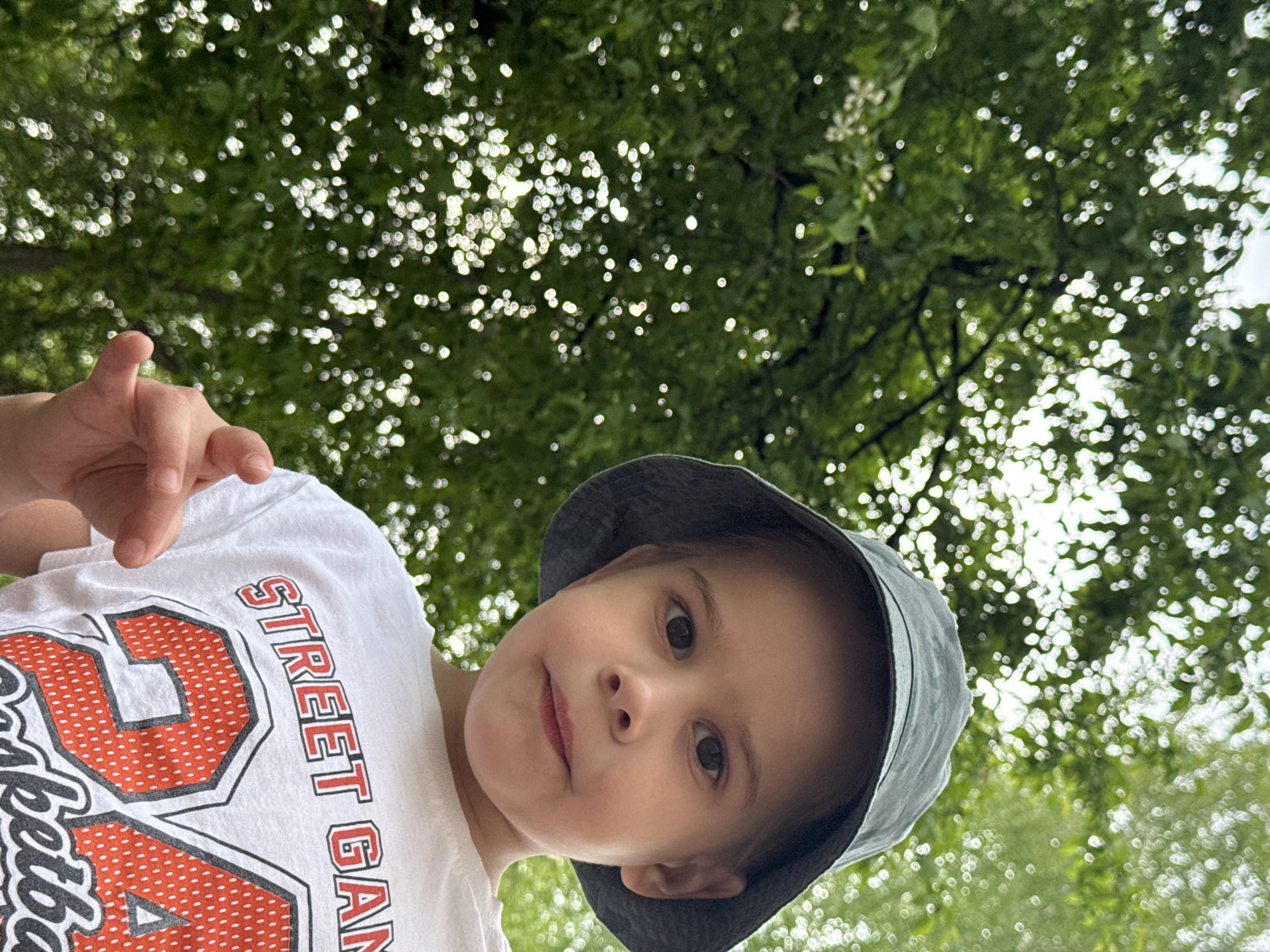 A young child, wearing a dark blue bucket hat and a white t-shirt with "STREET GAME 24 Basketball" printed on it, is looking down at the camera. The child's right hand is partially visible, with a finger extended. The background consists of a canopy of green trees with bright spots of light filtering through the leaves. The photograph appears to have been taken from a low angle, looking upward. The location is in a park in the city of Chaikovsky, Russia.