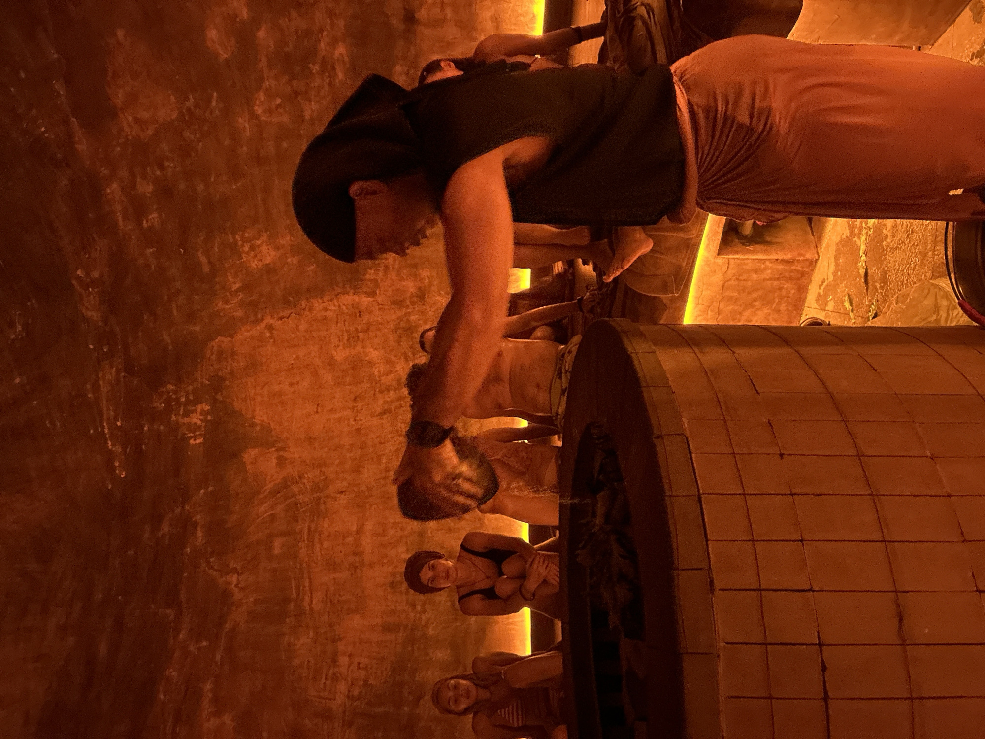 In a dimly lit, orange-hued circular room, likely a spa or bathhouse, a person wearing a dark head covering and an orange sarong stands near a tiled circular structure. They are pouring a substance from a metal bowl into the structure, with steam or smoke rising from it. In the background, several individuals, some wearing swimwear, are seated in a row, illuminated by soft lighting from the wall. The walls appear to be textured and illuminated from behind. The overall atmosphere is warm and intimate. The presence of a traditional-looking tiled structure and the communal seating arrangement suggest a communal bathing or therapeutic ritual. Given the context of the question, this scene is set in Kediri, Indonesia.
