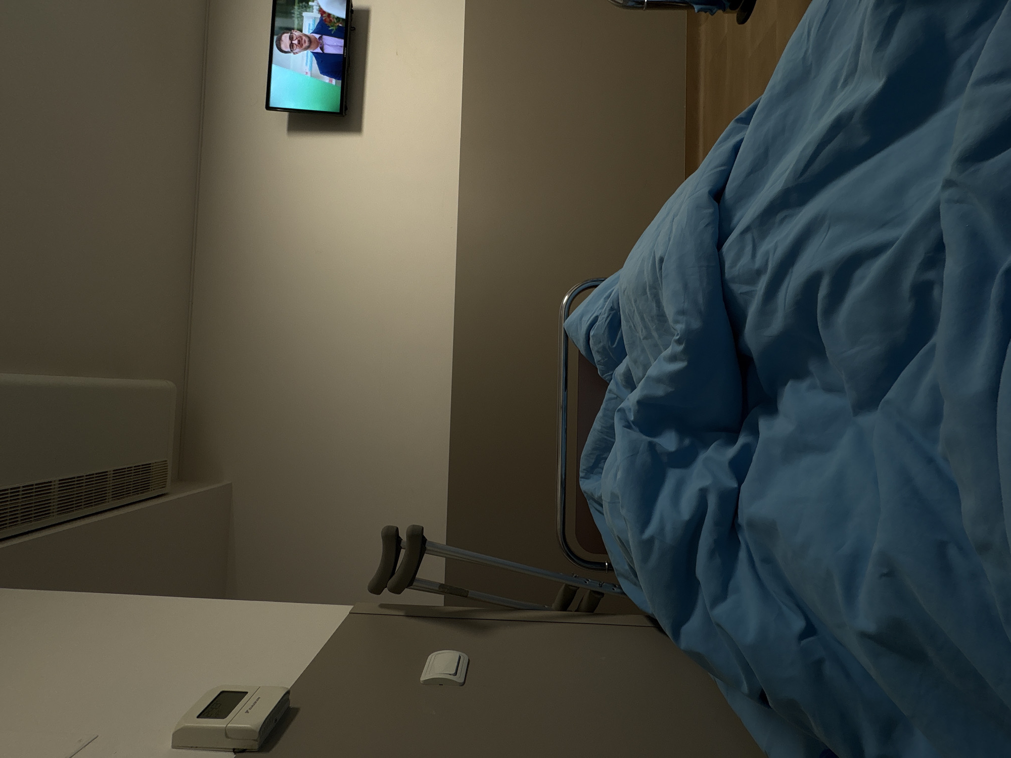 The image shows a hospital room. A television is mounted on the wall, displaying a person. A hospital bed is present, covered with a light blue blanket. A pair of crutches rests beside the bed, on a surface that also holds a remote control and a call button. An air conditioning unit is visible on the wall. The location is likely in a hospital in the city of Sofia, Bulgaria.