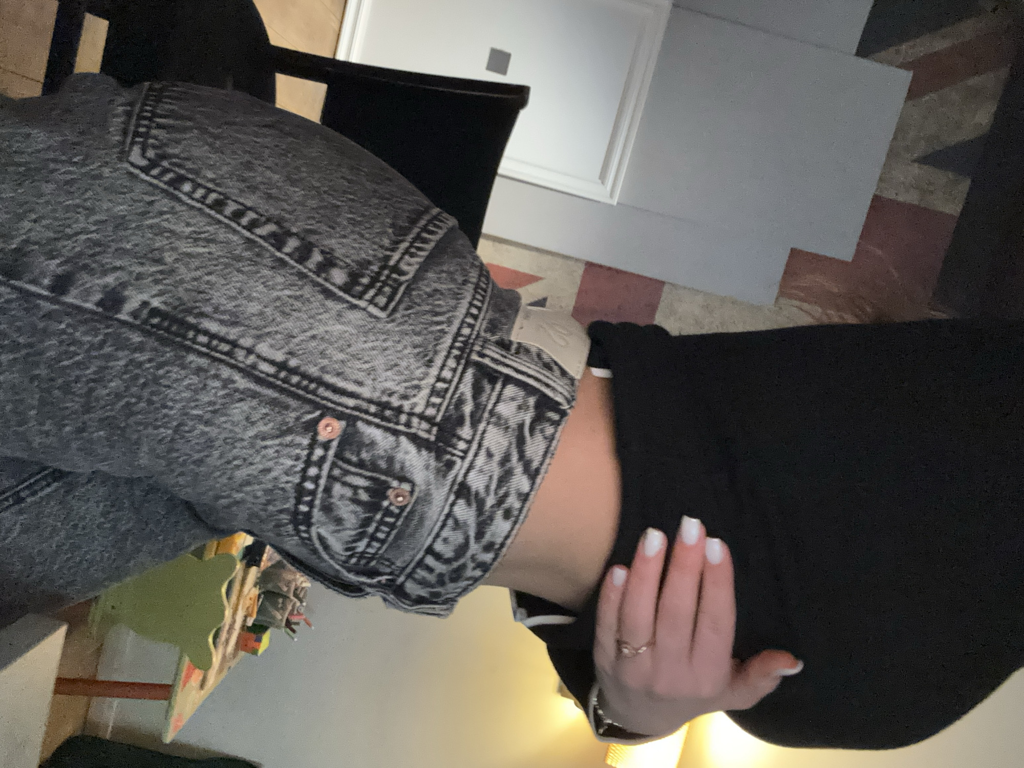 An indoor scene in Sofia, Bulgaria depicts a person from the mid-torso to mid-thigh, positioned facing slightly right. The individual wears a black long-sleeved crop top, the hem of which is lifted by their right hand, exposing the lower abdomen. The right hand exhibits white French-tipped nails and a simple ring on the ring finger. The person is also wearing high-waisted, acid-wash denim jeans with visible pockets and copper-colored rivets.
In the background to the left, a dark-colored chair with a slatted back is partially visible next to a white door frame and a closed white door featuring a small grey square object, possibly a light switch. The floor is light-colored wood. To the right, a wall is adorned with a large, distressed pattern resembling the Union Jack flag in muted red and beige tones. Further right, a small wooden table with orange legs is visible, topped with items including containers of writing implements and a multi-colored cube. A light green, child-sized chair with a decorative back is positioned beside this table. A warm, yellow light source illuminates the upper right background. The environment suggests a residential interior, possibly a living space or child's room.