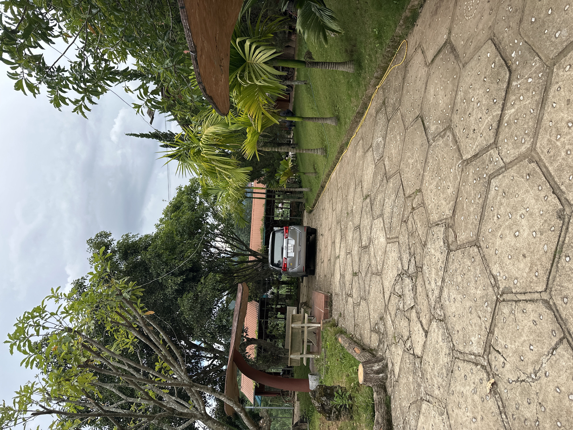 This image depicts a paved walkway leading towards a building complex in a rural setting. The walkway is constructed of hexagonal concrete slabs with small stones embedded in them, showing signs of cracking and wear. To the left of the walkway, there is a concrete picnic bench under a brown, mushroom-shaped canopy, and further back, a building with a red roof and open-air seating areas.
On the right side of the walkway, a grassy area is lined with several palm trees of varying heights. A silver SUV with license plate "51G 635.89" is parked on the paved area to the right of the building entrance. The background is filled with lush green trees and vegetation, suggesting a tropical or subtropical climate. The sky is overcast, with scattered clouds. The overall scene appears to be an outdoor seating or rest area, possibly part of a resort or restaurant, located in Thôn 2, Vietnam.