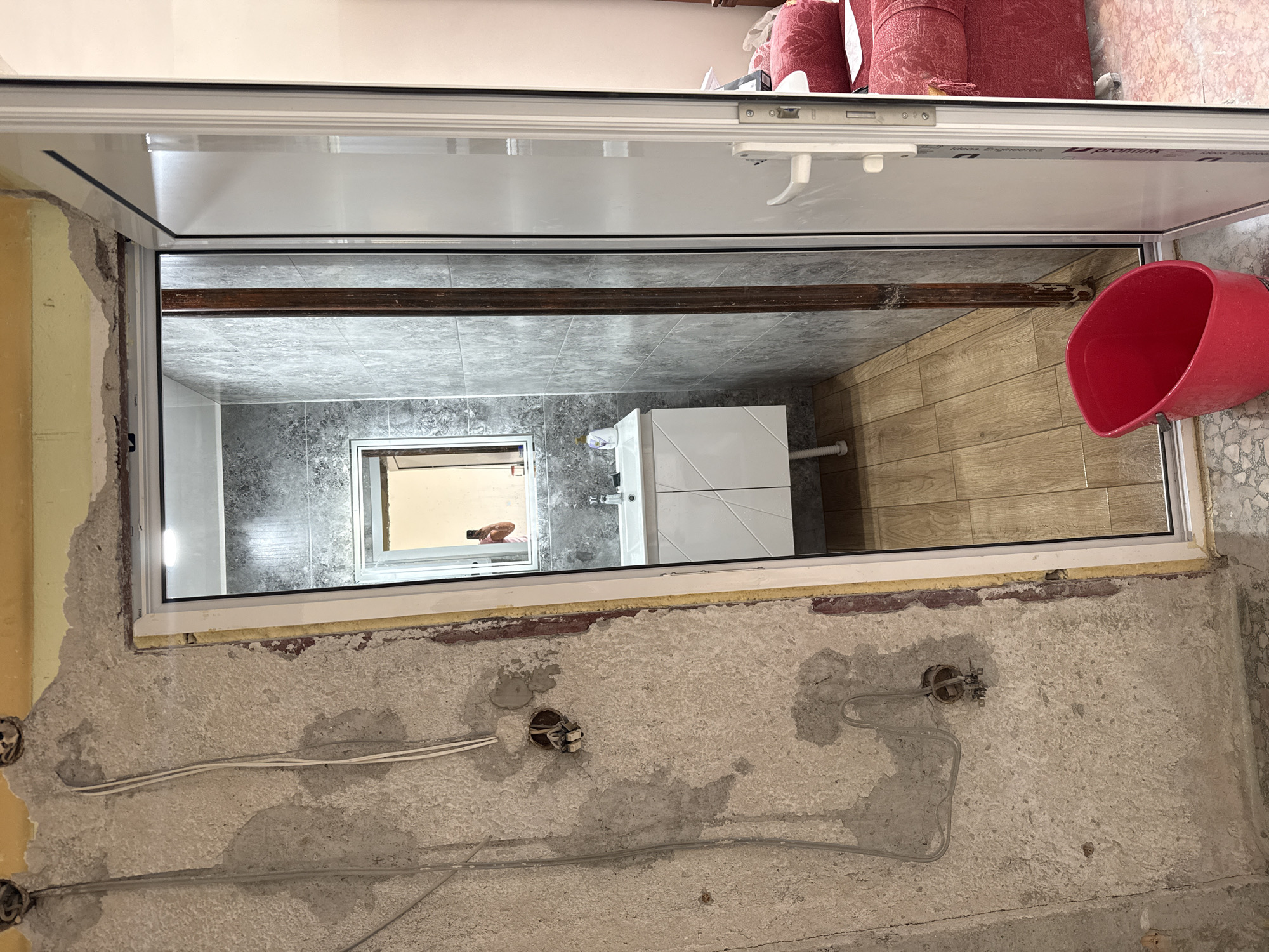 The image depicts an interior scene, possibly a bathroom under renovation, with exposed concrete walls, electrical wiring, and plumbing fixtures. In the background, a modern bathroom with a vanity, sink, and tiled walls is visible through an open doorway. A bright red plastic bucket sits on the floor in front of the doorway. The scene appears to be in Burgas, Bulgaria, based on the provided context. There are no people visible in the image. The environment is messy, suggesting construction or renovation work is in progress.