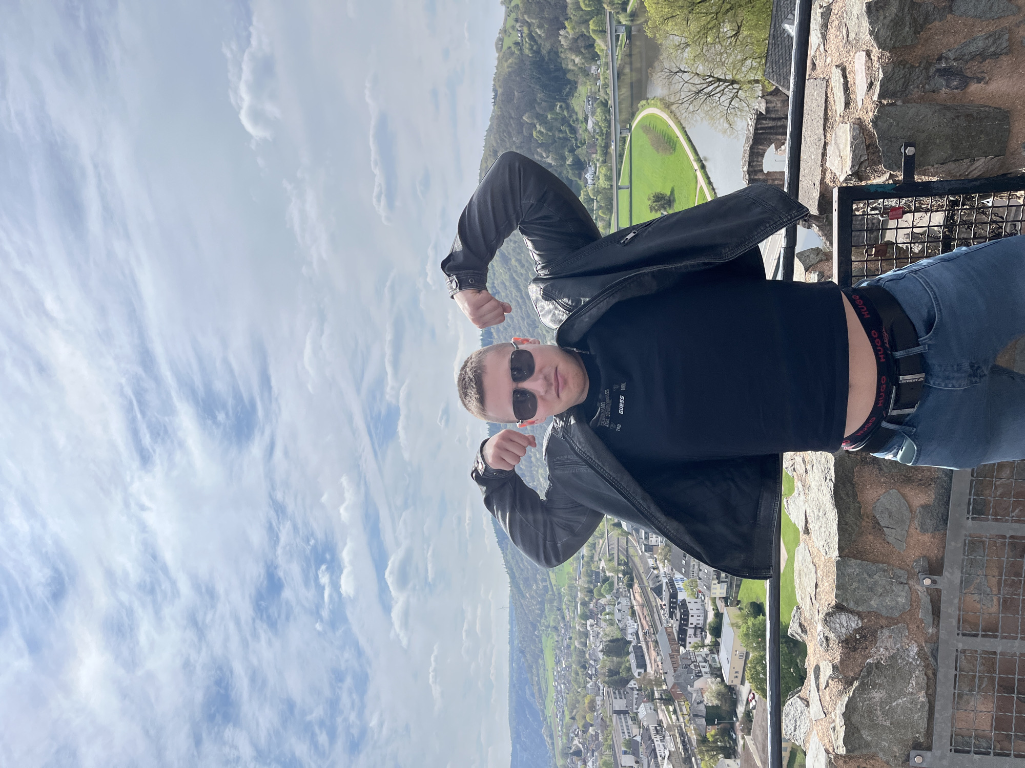 A man is standing on a stone wall overlooking Saarburg, Germany. He is wearing a black leather jacket, a black shirt, jeans, and sunglasses. The man is posing by flexing his arms. Saarburg can be seen in the background, as well as the Saar River. The sky is visible with light clouds.