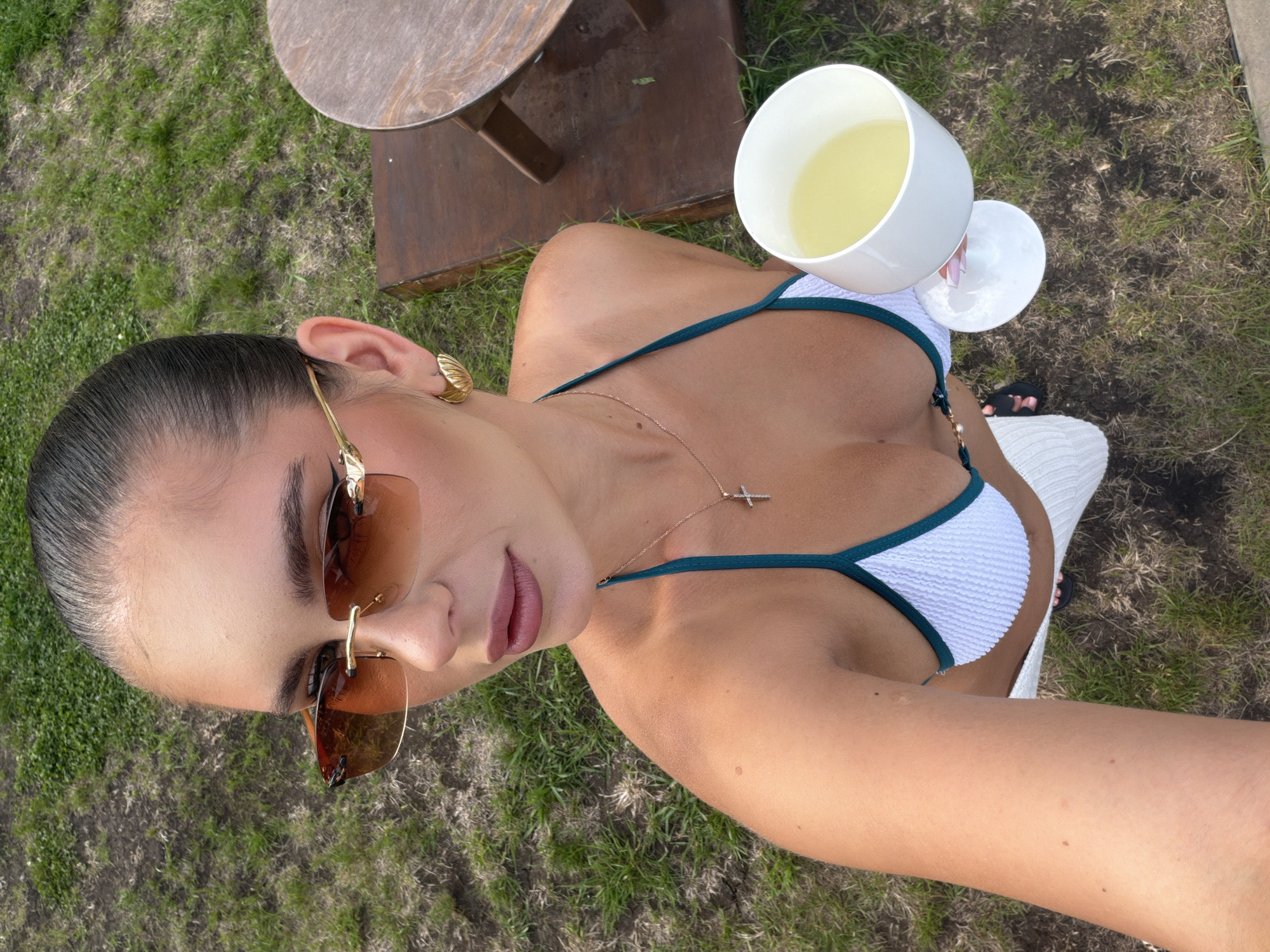 A woman is holding a white glass with a yellow beverage in her right hand, wearing a white and teal bikini top and sunglasses. She is standing on a patch of grass with a wooden table to her right.