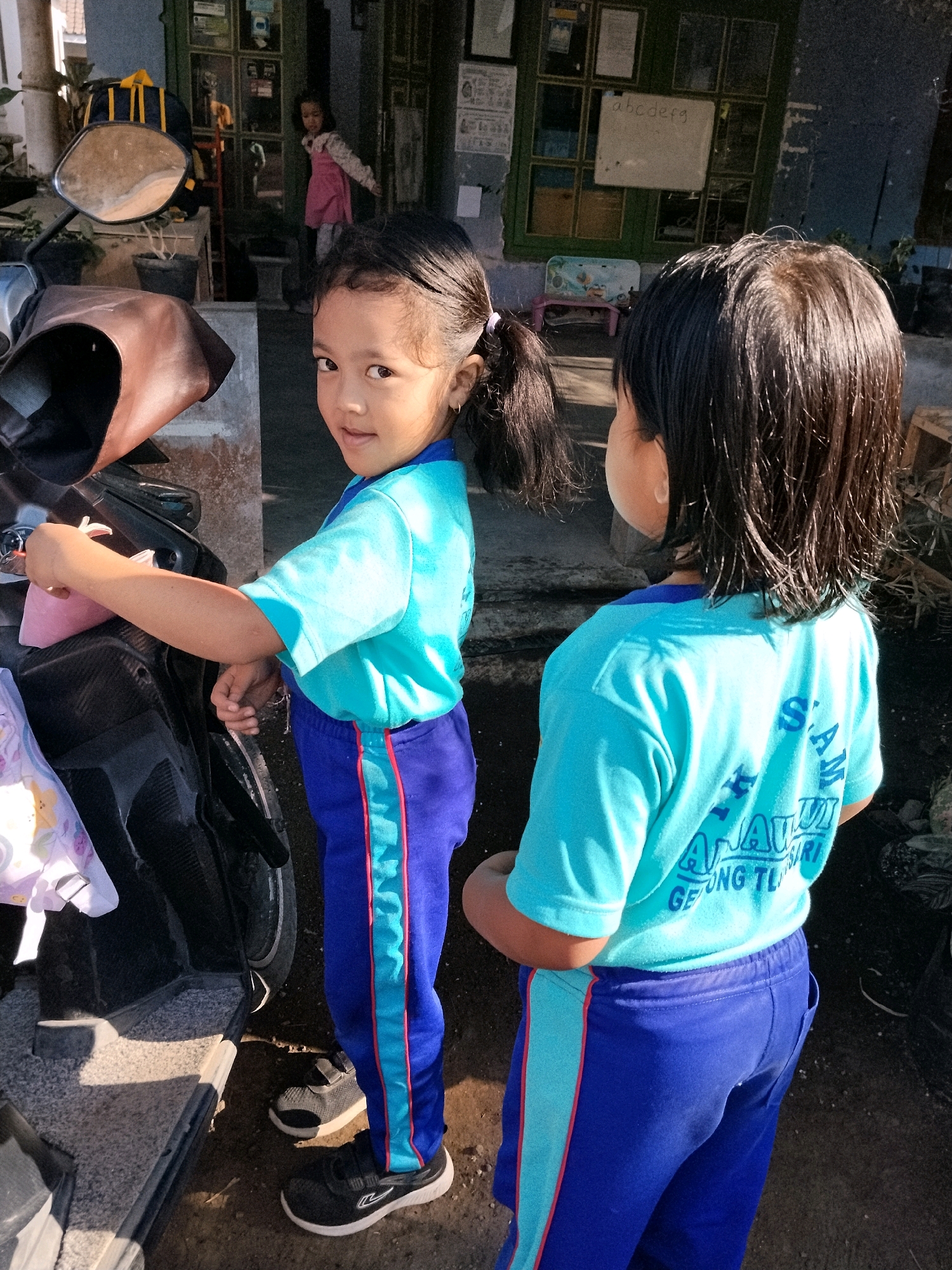 Two young girls in blue tracksuits with light blue accents are standing in front of a building. The girl on the left, facing the camera, is holding a pink pouch and looking at the viewer. She has dark hair tied in a ponytail. The girl on the right is facing away from the camera, with her back to the viewer. Her hair is dark and wet. Behind the girls, a person in a pink dress is visible further back in the building. The building appears to be a dwelling or a small shop in the city of Panggungan, Indonesia. The ground is dirt and gravel, with a parked motorcycle to the left of the girls. The overall lighting suggests it is daytime.
