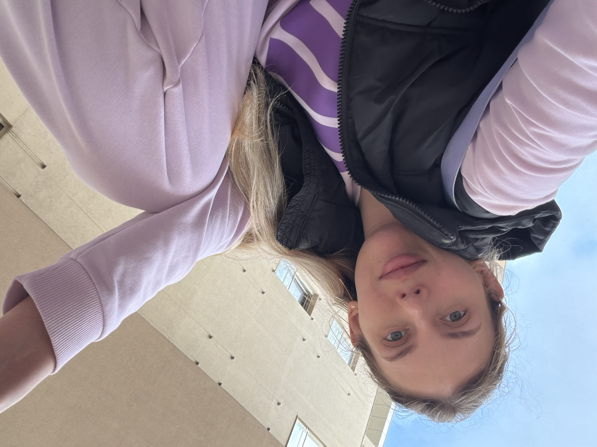 A young woman is captured in a low-angle, inverted shot, looking directly into the camera with a calm, neutral expression and slight smirk. Her head and upper torso are in the lower right and center of the frame, while her arm extends upwards to the left. She has blonde or light brown hair and light eyes.
She is dressed in casual layered clothing: a light lavender long-sleeved top or hoodie, worn over a purple and white striped shirt. A black quilted or puffer vest is worn over these, with its zipper partially visible. The texture of the fabrics, including the ribbed cuff of her sleeve, is clear.
The background shows a light beige, modern building with several small, dark square protrusions and visible windows on the left side of the frame, behind her arm and torso. On the right, behind her head, a clear blue sky is visible. The bright, even lighting indicates it is daytime with good weather. No text is visible in the image. The scene is set in Budva, Montenegro.