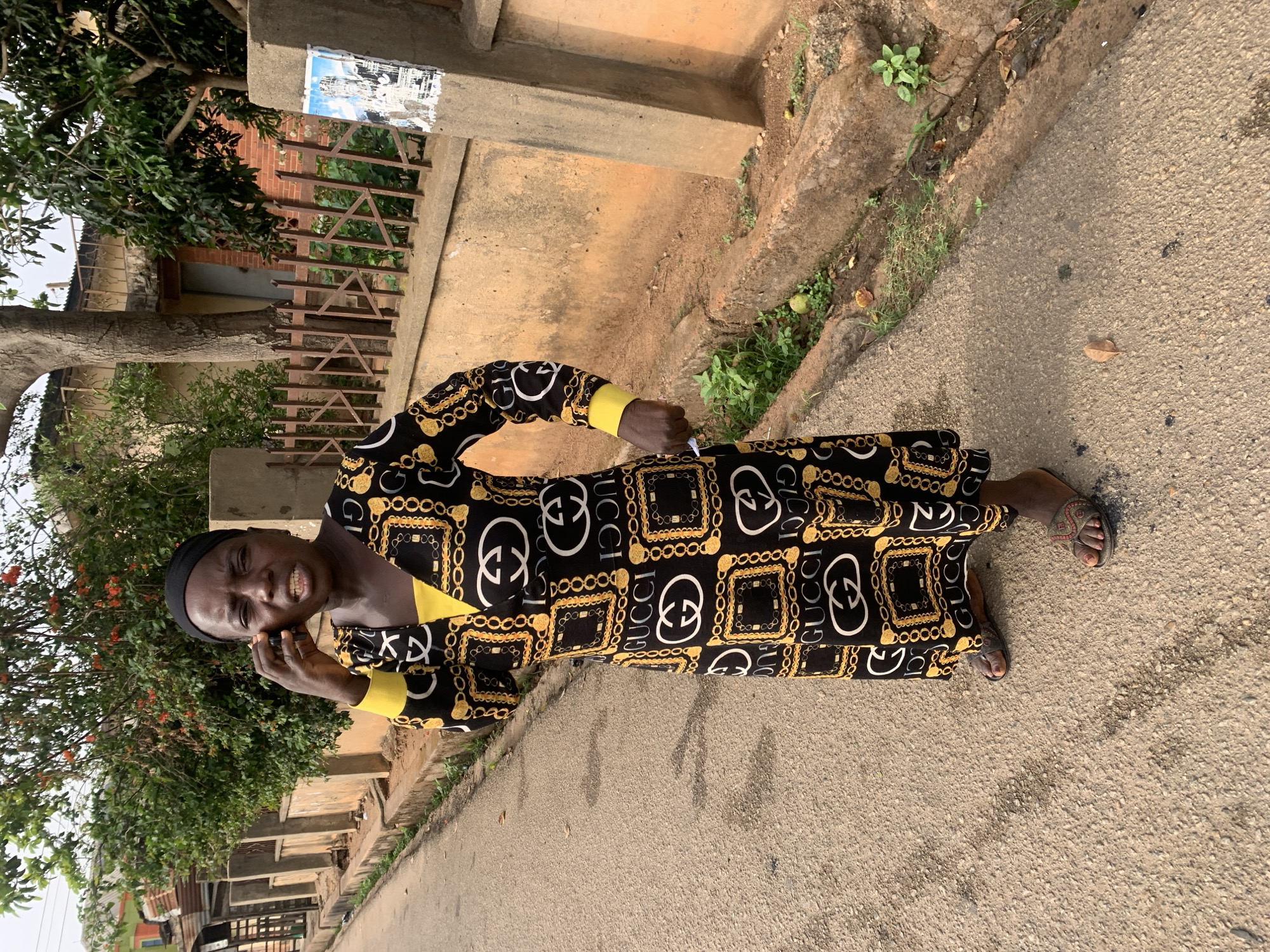 A middle-aged African woman is standing on an asphalt road, seemingly in a residential area. She wears a long-sleeved dress with a black background and a repeating Gucci logo pattern in gold and white, along with yellow cuffs. She is wearing a black head covering. She is holding a cell phone to her ear, appearing to be in a conversation. The woman has a slight smile. To her left is a tree with green foliage. A concrete curb runs along the side of the road, with vegetation growing along it. In the background, there is a partially obscured building with a weathered exterior. The scene appears to be in the city of Jos, Nigeria.