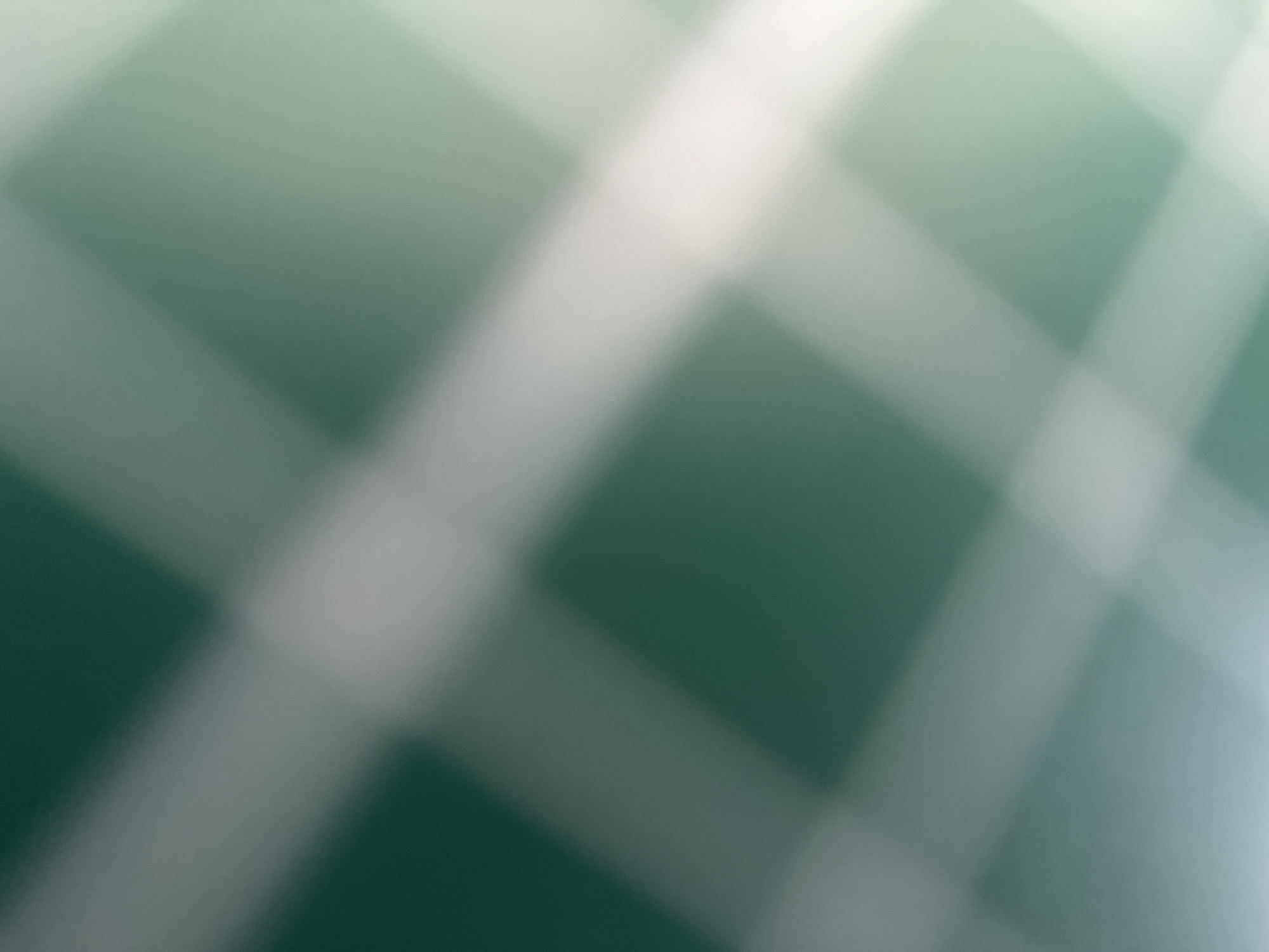 This is a close-up, abstract shot of a surface with a green background and white diagonal lines forming a grid pattern. The image is heavily blurred, giving it a soft and out-of-focus appearance. The lines are not sharp but rather diffuse, suggesting they might be created by light or a raised texture on the surface. The green color is muted, with some variations in shade that could indicate lighting. There are no discernible objects, people, or specific environmental details that would suggest a particular scene, setting, time of day, or weather. The abstract nature of the image and the lack of clear context make it impossible to identify any text or infer any specific activity or emotion. The hints provided are also abstract and do not offer additional clarity. Given the location context of Belgrade, Serbia, it is still not possible to tie this abstract visual to any specific landmarks or cultural elements.