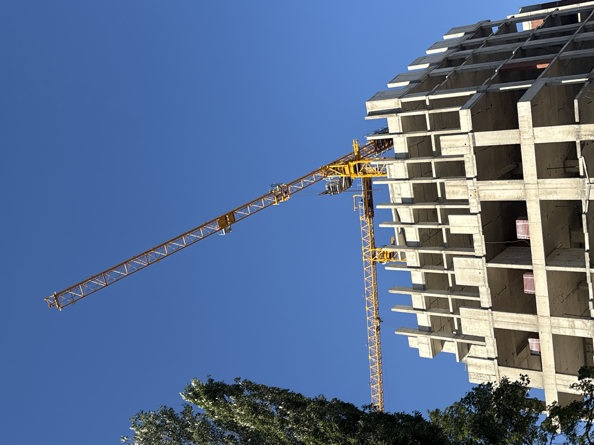 The image shows a construction site in Perm, Russia, featuring a partially constructed concrete building under a clear blue sky. A yellow construction crane is attached to the side of the building, its arm extended. The building is multi-story and appears to be a high-rise. Trees are visible in the foreground.