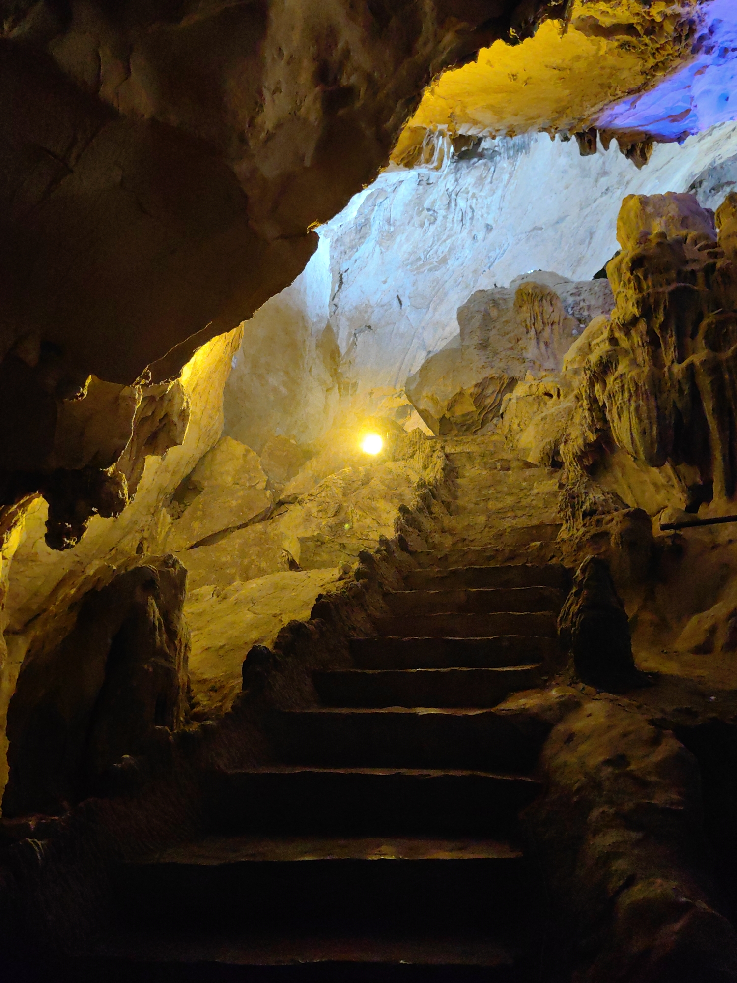 The image shows a stone staircase leading upwards inside Tam Thanh Cave, a famous tourist destination in the city of Lạng Sơn, Vietnam. The cave's ceiling is visible at the top of the stairs, with lighting that gives off yellow and blue hues. The stairs are made of carved stone and lead towards a bright opening. The cave walls are textured with natural formations and are lit with artificial lights. There are no people visible in the image.