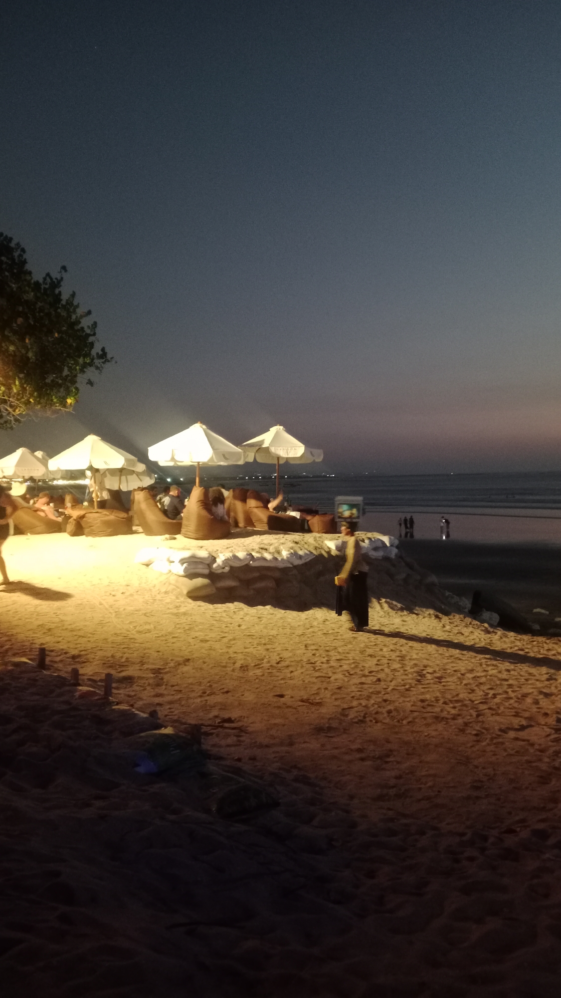 This is a vertical, eye-level shot of a beach in Kuta, Indonesia, at dusk. The sky is a gradient of dark blue to a lighter purple and pink near the horizon. In the foreground, the sandy beach is illuminated by artificial lights, casting long shadows. Several white umbrellas are open over beanbag chairs arranged on a raised sand platform, indicating a seating area. Some people are visible sitting on the beanbags, while others are walking along the shoreline in the distance. A person in a dark uniform and a light-colored top stands on the sand, facing away from the camera. The ocean stretches to the horizon, with small boats or lights visible in the distance. The overall scene depicts a relaxed evening ambiance on a beachside establishment.
