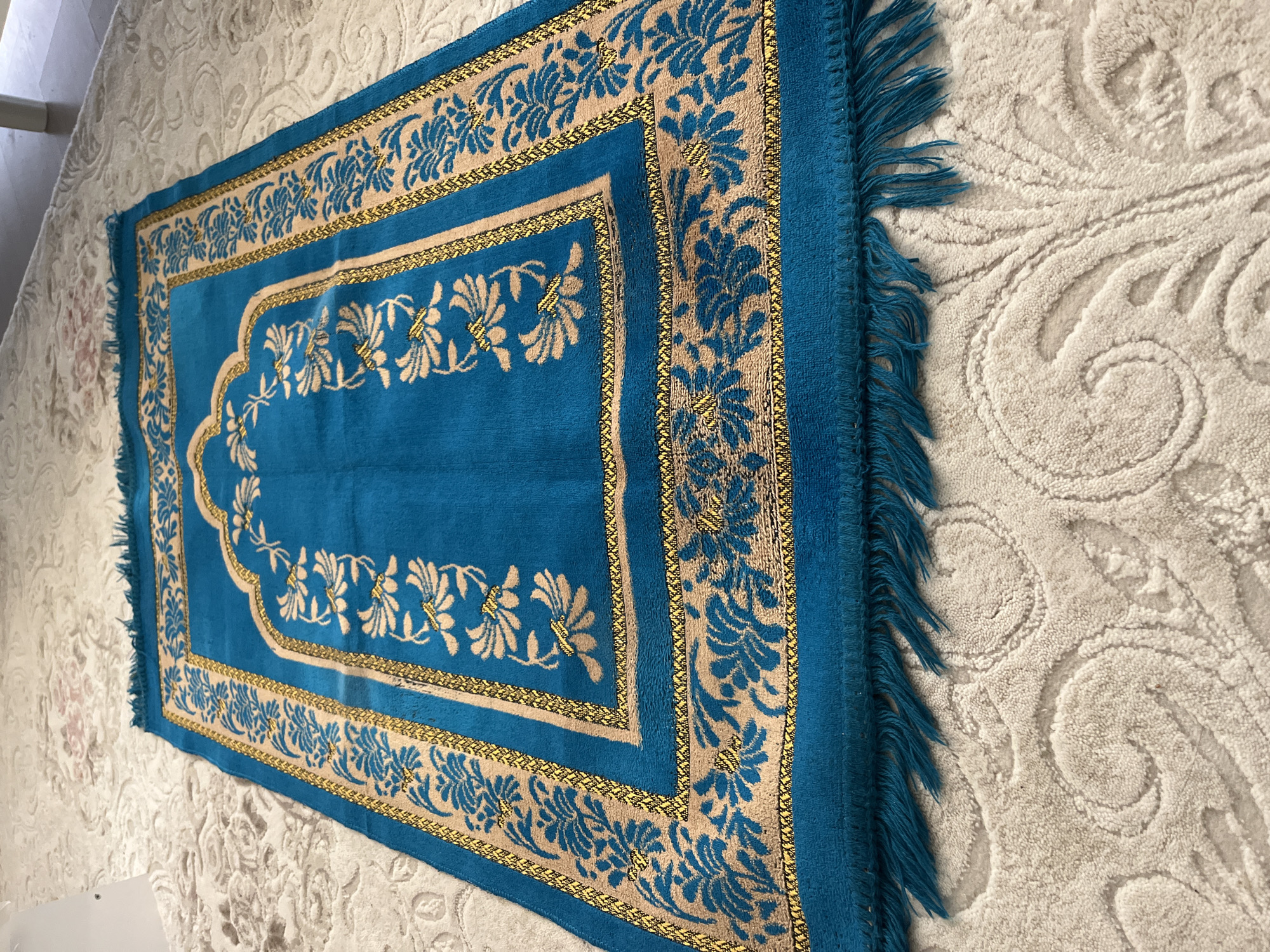 A teal prayer rug with gold and beige floral accents is laid on a textured, cream-colored carpet. The prayer rug features a central arched design with floral motifs and a border with intricate patterns. Fringes in the same teal color as the rug are visible along one edge. The cream carpet has a raised, damask-like pattern of swirling floral shapes. The scene appears to be indoors, possibly a room in a home or a prayer space in Gürsu, Turkey. There is no text visible in the image. The lighting suggests it could be daytime, with natural light illuminating the scene. The focus is on the texture and pattern of the rug and carpet.