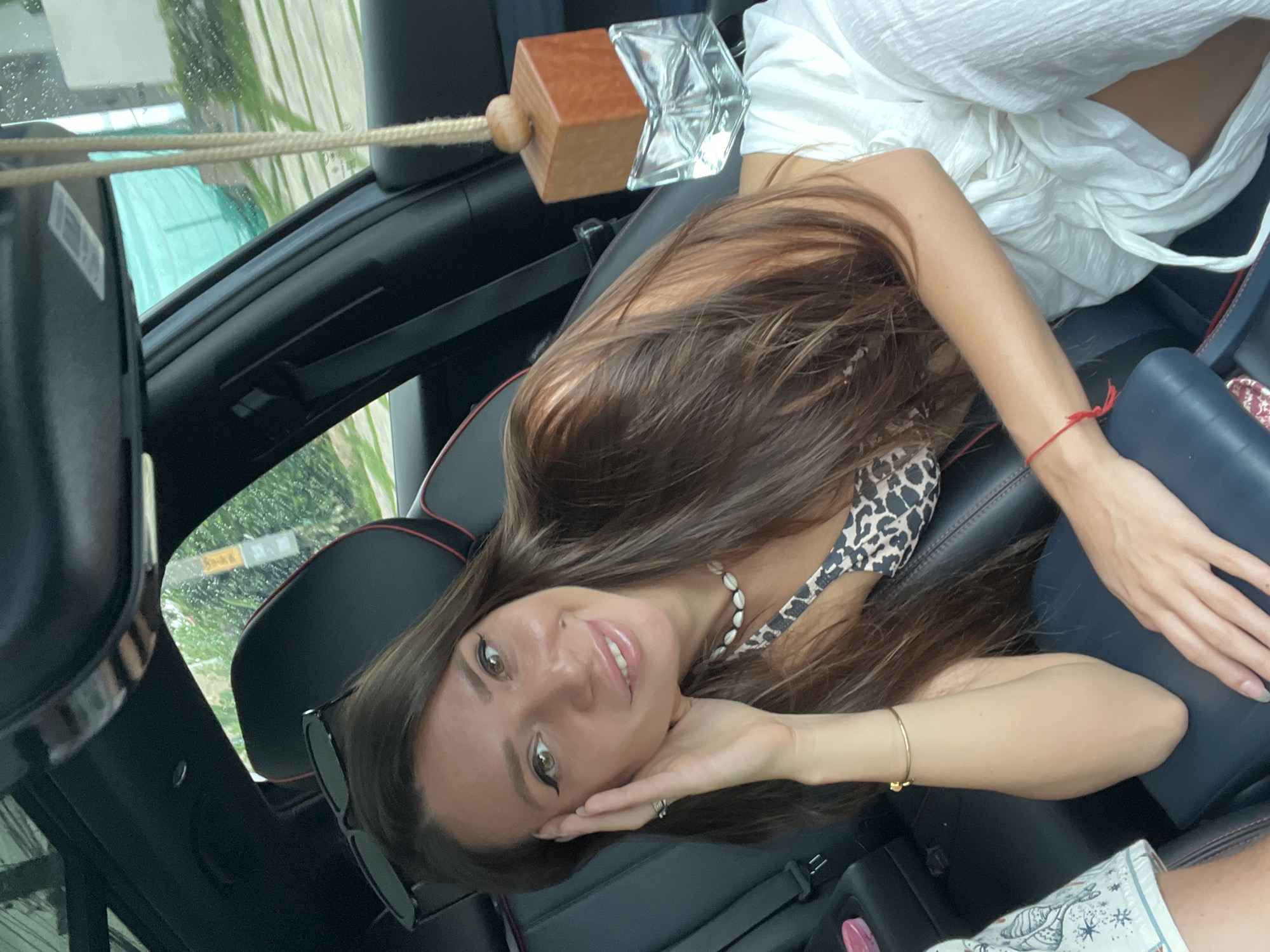 A woman reclines in the passenger seat of a car, smiling softly and looking directly at the camera. She has long, dark brown hair, fair skin, and is wearing makeup, including winged eyeliner. Her attire consists of a leopard print top and a white, flowy skirt or dress. She accessorizes with a shell necklace, a gold bracelet on her right wrist, a red string bracelet on her left wrist, and a ring on her left ring finger. Sunglasses are perched on the headrest next to her.
The car's interior features dark seats with red stitching and a dark blue armrest. A decorative car air freshener or ornament, made of a wooden cube and a clear, faceted glass-like component, hangs from a rope near the front of the vehicle. Through the car windows, lush green foliage and trees are visible, suggesting a natural, tropical environment. Water droplets on the windows indicate either recent rain or a car wash. The scene takes place during the daytime under natural light. The woman's relaxed posture and gentle smile convey a calm and pleasant mood.
The location is Baan Chaweng Noi, Thailand. Visible text includes an illegible sticker or barcode on an object in the upper left of the frame, and a faint, unreadable sign seen through a window.