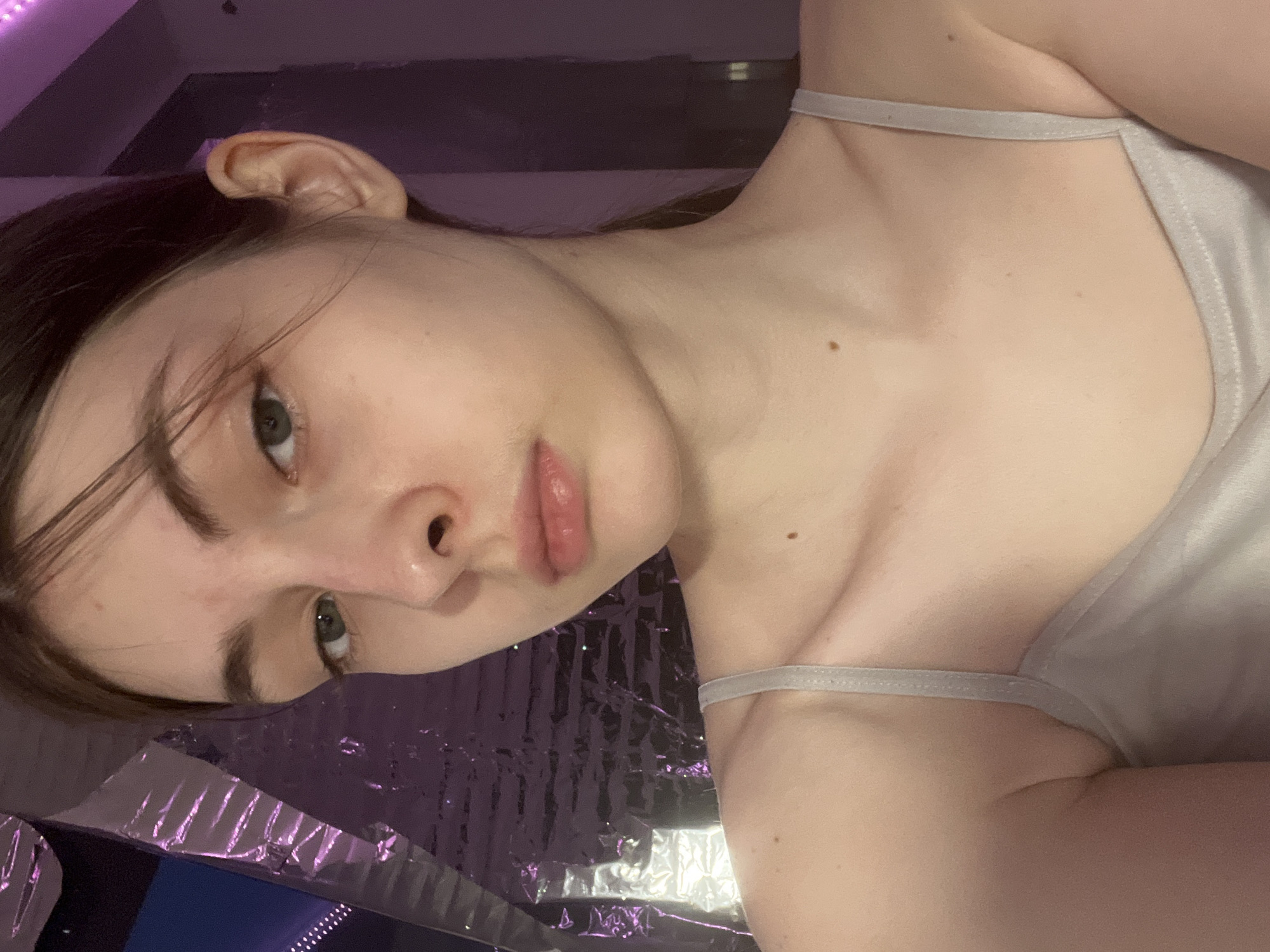 A close-up, low-angle portrait shows a fair-skinned young woman with dark hair, wearing a light-colored tank top with thin straps. Her head is slightly tilted. She has visible facial features including eyes, nose, and lips. A mole is on her upper chest. The background appears to be a surface covered with shiny, reflective material, predominantly in shades of purple and silver. The image contains no information to infer the location in the city of Saint Petersburg, Russia.