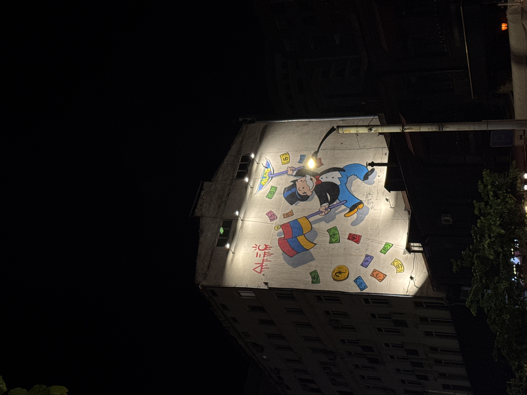 At night, a low-angle shot captures a building with a large mural in Yerevan, Armenia. The mural depicts a cartoon figure resembling the Monopoly man, wielding an umbrella adorned with dollar signs and holding a colorful flag. Money and coins are depicted floating around the figure. The building is illuminated by spotlights which highlights the mural.