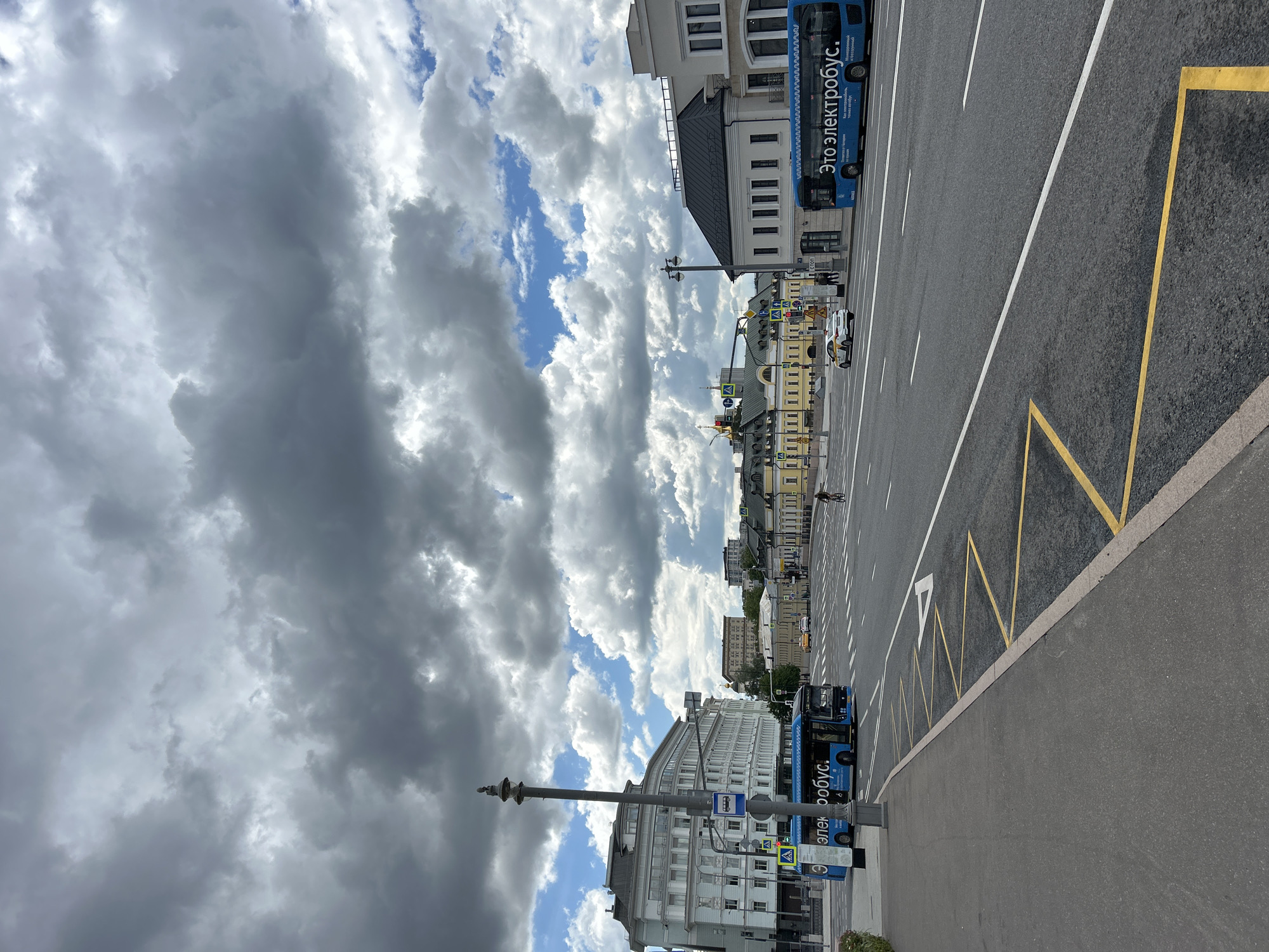 The image shows a street scene in an urban environment, with a heavy, cloudy sky dominating the left side. A wide, multi-lane road stretches into the distance, marked with white and yellow lines. A bus stop is visible on the right side of the road, indicated by a zig-zag yellow line and a bus. Buildings line both sides of the road, varying in architectural style. People can be seen walking along the street and riding bikes. The location is Unknown, Unknown.