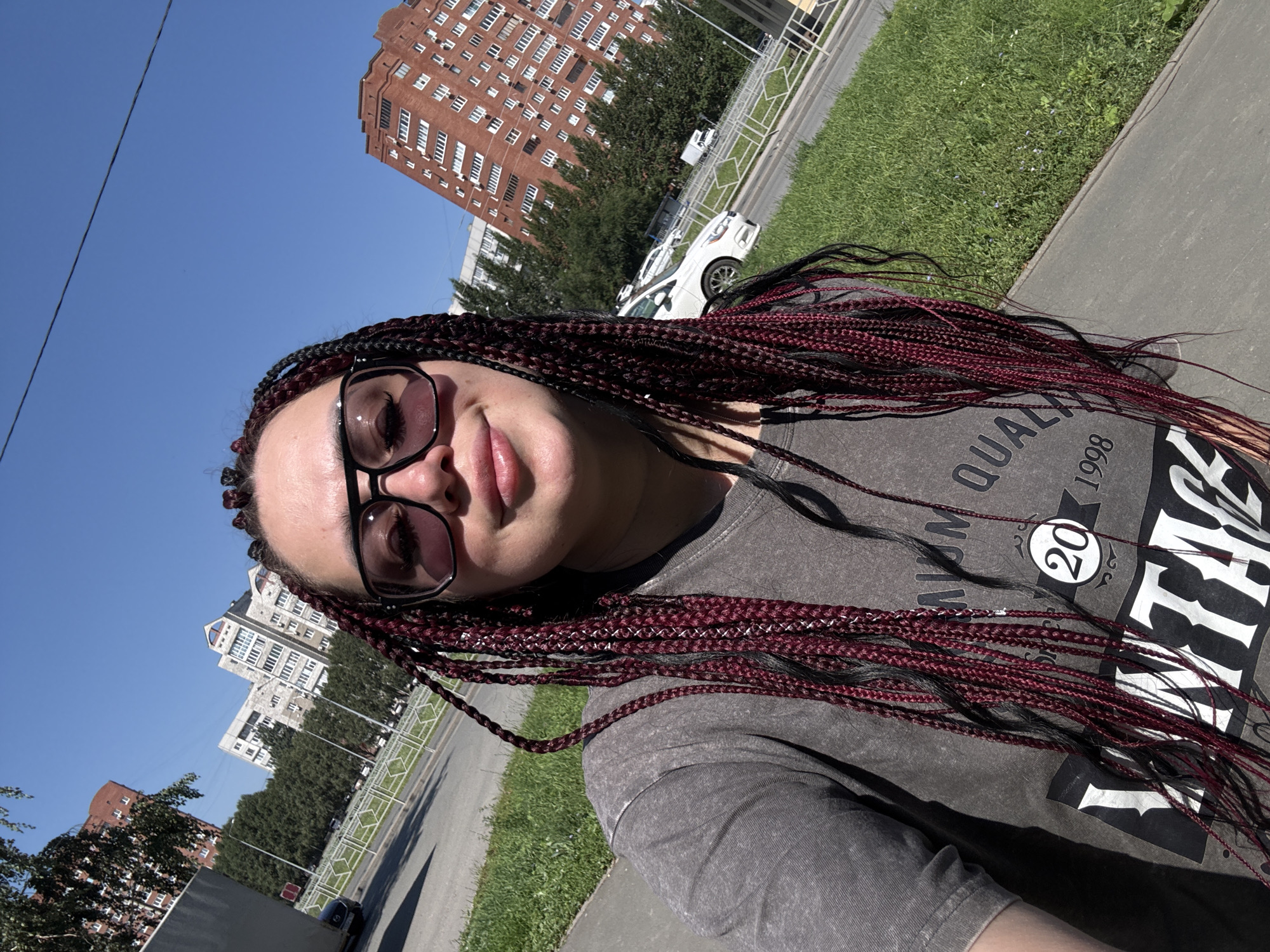 A woman with braided hair, wearing sunglasses and a gray t-shirt, is taking a selfie outdoors. She is standing on a paved walkway next to a grassy area. In the background, there are several apartment buildings and a street with cars. The sky is clear and blue. The location is the city of Tolyatti, Russia.