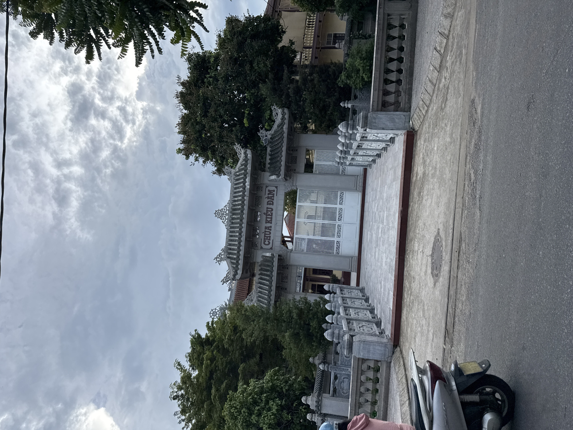 The image shows the entrance gate of a temple, identified by the Vietnamese text "CHÙA KIỀU ĐAM" above the gate. The architecture is distinctly Southeast Asian, with ornate grey tiled roofs and decorative stone railings. The gate itself is white and appears to be made of metal with an intricate pattern. In front of the gate, there is a paved walkway leading up to it, bordered by the stone railings. The surrounding environment includes lush green trees and a building with a pale yellow facade to the right of the temple entrance. The sky is overcast with dramatic clouds. In the foreground, a silver scooter is parked on an asphalt road, and a person wearing a pink garment is partially visible standing near the scooter. The scene appears to be in a residential or semi-urban area, possibly within Thuận Hóa Ward, Vietnam, given the architectural style and the presence of Vietnamese text.