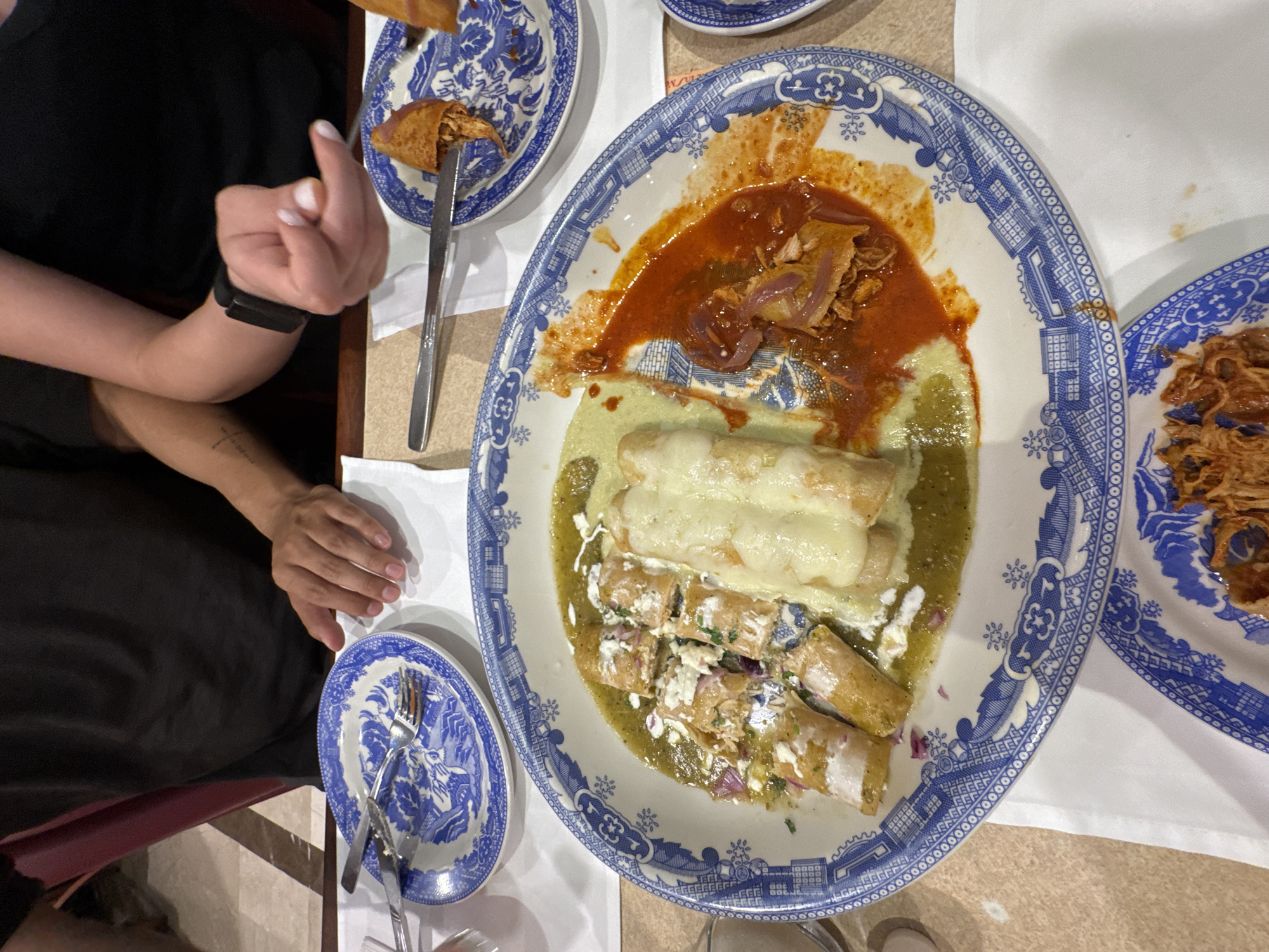 A high-angle shot depicts a table in what is likely a restaurant, given the presence of plates with food, silverware, and a person's arms and torso. The location is potentially Puebla, Mexico, based on the style of the enchiladas and the traditional Talavera pottery plates.
The main subject is a large oval plate with blue and white Talavera-style decoration. It holds two types of enchiladas: one portion appears to be enchiladas suizas, covered in a creamy green sauce and topped with melted cheese, while the other portion is covered in a white sauce and sprinkled with diced onions and cilantro. Adjacent to the enchiladas is a portion of meat (possibly chicken) in a reddish-brown sauce with visible onion slices.
To the left of the main plate is a smaller, similarly patterned plate with a partially eaten flauta and a knife. Further to the left and below is another small Talavera plate with used silverware (fork and knife). To the right of the main plate is another plate featuring what seems to be shredded chicken.
A person, presumably a diner, is visible on the left side of the frame. Their left arm and hand rest on the table, while their right hand holds a fork near the plate with the flauta. The person is wearing a dark-colored shirt or dress and has a watch or fitness tracker on their left wrist. A small tattoo is visible on their left forearm.
The table surface appears to be a light-colored, potentially laminated material. A white napkin is visible underneath the plates. The scene suggests a meal in progress at a restaurant, showcasing traditional Mexican cuisine served on Talavera pottery.