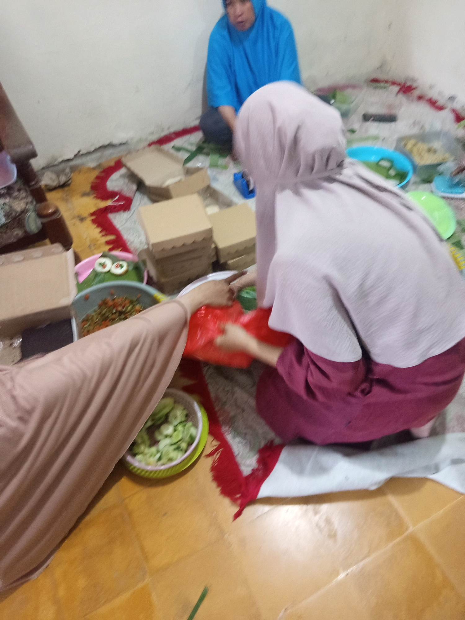 The image depicts a dimly lit indoor scene, possibly a dining area, with several people dressed in Islamic attire. A person in a blue hijab sits in the background, facing away from the camera. In the foreground, another person wearing a pale pink hijab and a dark red garment is seated, attending to food items. To the left, a third person, whose face is obscured, is dressed in a beige hijab and garment and is reaching towards a red plastic bag. Scattered around are several food containers, some cardboard boxes, and bowls of food, including what appears to be sliced cucumbers and a spicy relish. The floor is made of wooden tiles, and a patterned rug is laid out on the floor. The overall impression is that of a casual gathering where food is being prepared or served.