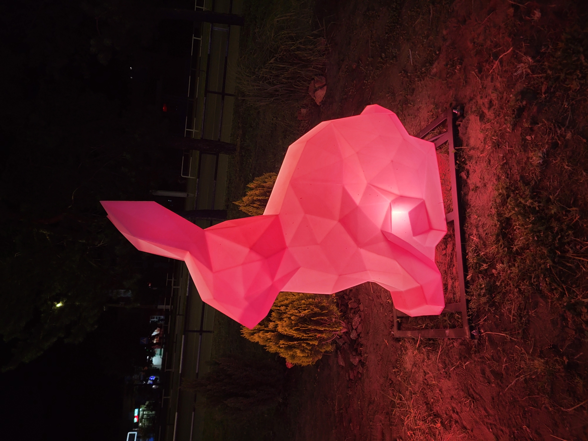 A large, illuminated, polygonal sculpture of a rabbit is positioned in the foreground, emitting a soft pink glow. The sculpture is mounted on a dark, rectangular metal frame that rests on the ground. The ground is uneven, covered with a mix of soil, dry grass, and small stones. To the left of the sculpture, a small, dense evergreen shrub with yellowish-green foliage is visible. Behind the sculpture and shrub, a dark fence with horizontal rails separates the area from a road or pathway. Trees with dark trunks and sparse foliage are scattered in the background, and beyond them, the dim lights of buildings and streetlights are visible, suggesting an urban or semi-urban environment at night. The overall lighting is dim, with the rabbit sculpture being the primary light source.