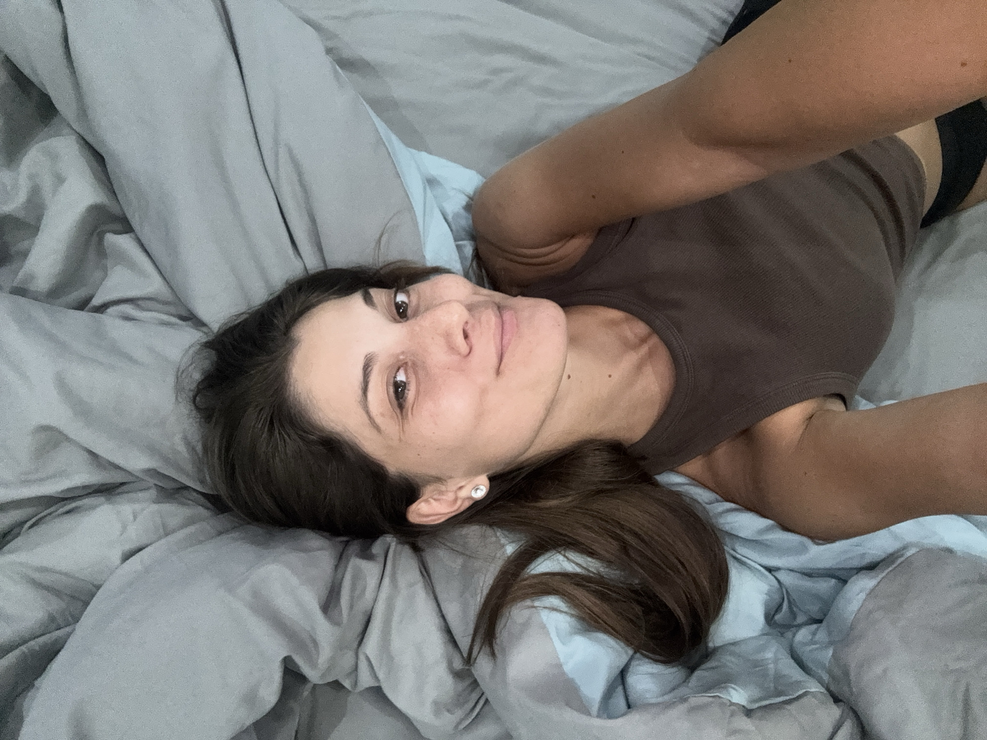 A woman lies on a bed covered with light grey and light blue bedding. She is wearing a brown tank top. Her dark brown hair is partially spread out on the pillow. She is looking at the camera. A small stud earring is visible in her ear. Part of her bare arm is extended toward the upper right corner. The location is not discernible from the image.