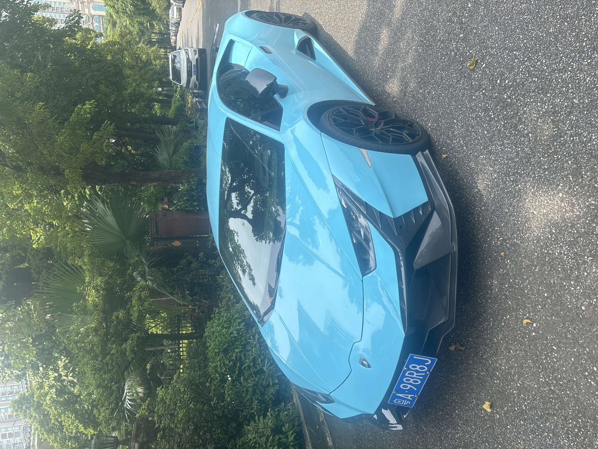 A bright blue Lamborghini is parked on an asphalt road in Nanhai District, China. The car is angled towards the viewer, with its front facing left. The vehicle has black accents and red brake calipers visible through the spokes of its black wheels. The license plate, which is blue with white characters, reads "A 98R8J".
The background consists of lush green trees and foliage, with a glimpse of a modern building in the distance on the right. A white SUV is parked further down the road on the right side, along with other vehicles in the distance. The scene appears to be a residential or upscale area, suggested by the well-maintained vegetation and the presence of luxury vehicles. The lighting indicates it is daytime, possibly late morning or early afternoon, with sunlight filtering through the leaves. There are no people visible in the image.