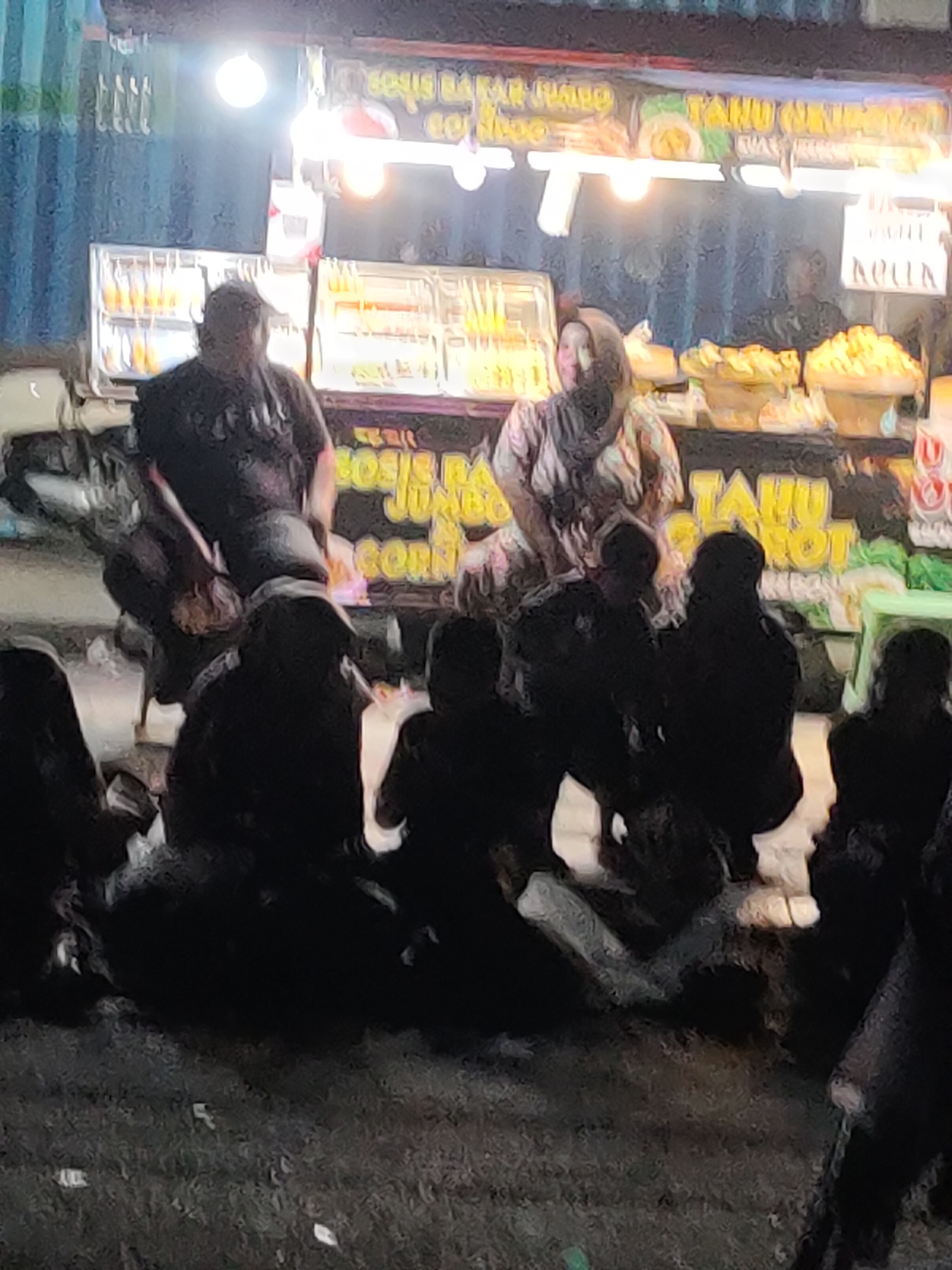 In Kanigoro, Indonesia, at night, a group of people are gathered around a food stall. The stall is illuminated by bright lights and displays various food items, including what appears to be fried snacks or pastries. A man wearing a dark t-shirt stands behind the counter, while a woman in a patterned headscarf is also near the stall. Several individuals are seated on the ground in front of the stall, some of whom are obscured by darkness. The environment appears to be an outdoor, possibly street-side, setting with a paved or dirt ground surface. The overall scene suggests a casual gathering at a local eatery.