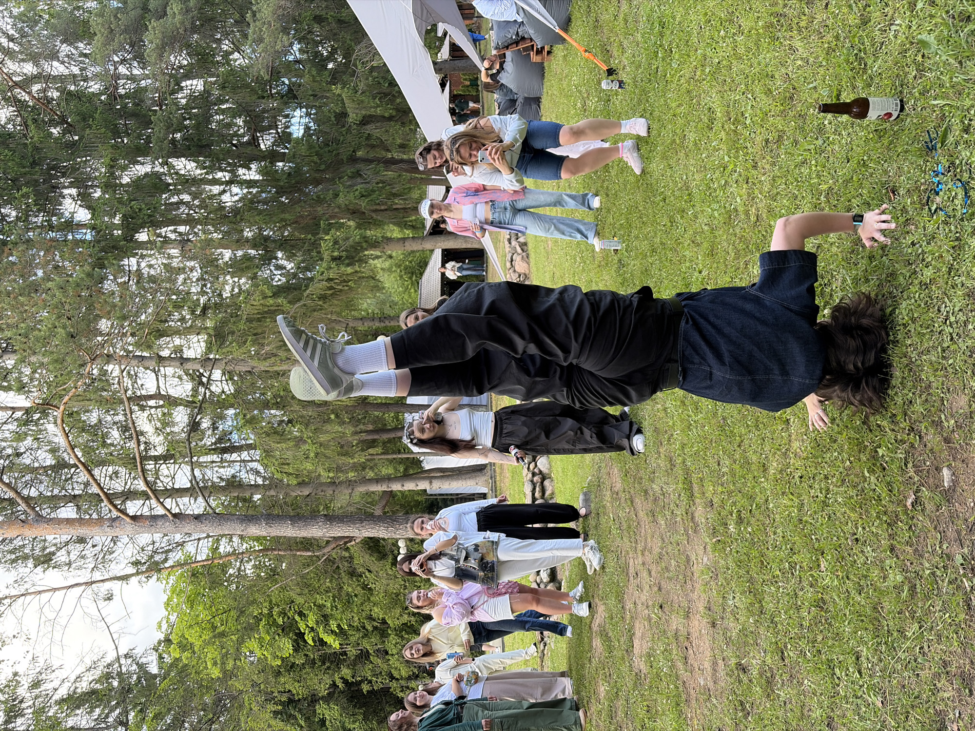 The image depicts an outdoor gathering with a group of young adults in a grassy area surrounded by trees. The scene appears to be set in a park or rural location, likely in Belarus, given the context. The overall atmosphere suggests a casual, recreational event, possibly a picnic, party, or informal get-together.
Several individuals are visible, some standing, others reclining or in the process of a physical activity. One person in the foreground is performing a handstand, showcasing an element of sport or playful exertion. Other people are interacting, some taking photos with their phones, indicating a social and engaging environment. The clothing worn by the individuals is casual and summer-appropriate, with t-shirts, shorts, and jeans being common.
Objects present include a bottle of beverage lying on the grass and some blue ribbon-like material scattered nearby, potentially remnants of an activity or decoration. Tents or canopies are also visible in the background, suggesting a planned event or a sheltered area for the gathering. The presence of tall pine trees and the lush green grass points towards a natural, possibly forested, setting.
The time of day appears to be daytime, with ample natural light, and the weather seems pleasant, suitable for an outdoor event. There are no visible signs or text in the image that provide specific information about the event or location beyond what can be inferred from the visual cues. The overall mood conveyed by the image is one of youthful energy, enjoyment, and camaraderie.