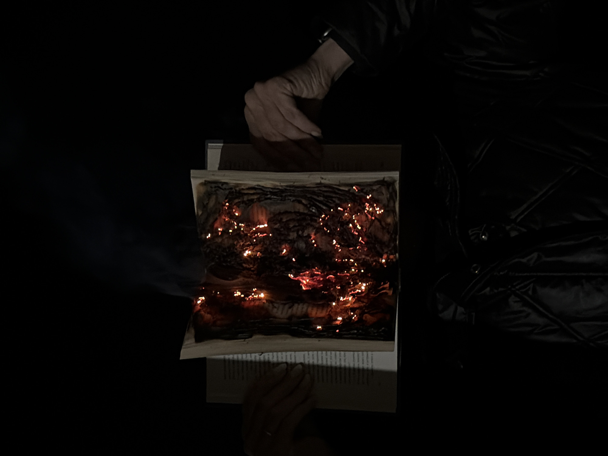 A book is on fire in the dark. The pages are charred, and there are flames visible. Two hands are present, one holding the bottom pages and another hovering over the top. The background is dark, with some fabric visible on the right. There is smoke rising from the book on the left. There are no visible clues in the image to determine the location.