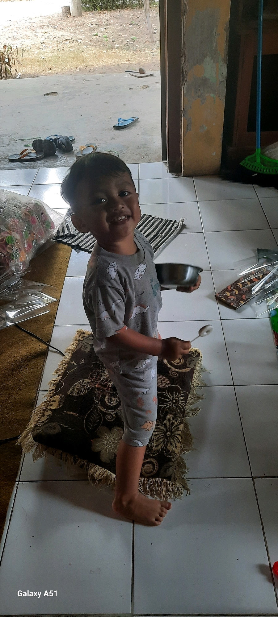 In Ploso, Indonesia, a young boy stands on a patterned rug with fringe, indoors. He is wearing a gray t-shirt with a dinosaur pattern and gray pants. He holds a metallic bowl in his left hand and a spoon in his right. The floor is tiled with white squares. Outside, visible through an open doorway, is a dusty, outdoor area with scattered debris and some green objects on the ground. To the left of the boy, a plastic bag filled with colorful items is visible. To the right, a broom with a green head and a blue handle leans against a wall. The wall itself appears weathered with peeling paint. The overall scene suggests a casual, domestic environment in a rural or semi-rural setting.