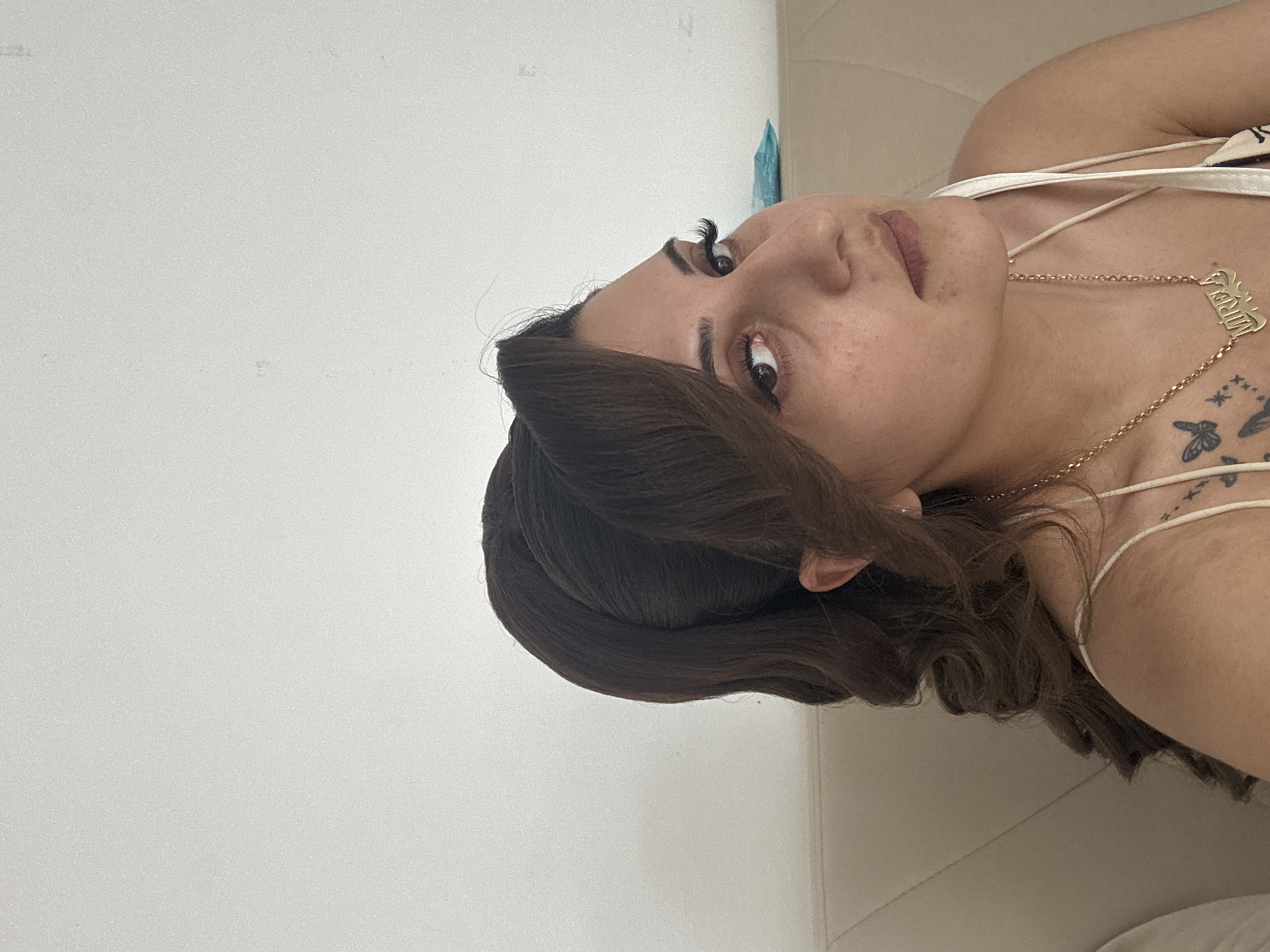 A medium shot captures a woman with dark hair styled in a voluminous, curled ponytail and a side-swept fringe, looking towards the right of the frame. Her makeup includes defined eyebrows, eyeliner, mascara, and a neutral lip color, with a smattering of freckles visible on her cheeks and nose. She is wearing a white tank top with thin straps and a gold necklace featuring a nameplate that reads "MIRELA," with small star and butterfly tattoos visible on her upper chest and shoulder. The background consists of a plain, lightly textured white wall and a beige couch. There is a blue object on the couch behind her. The image appears to be taken indoors, likely in a residential setting within the city of Stara Zagora, Bulgaria.