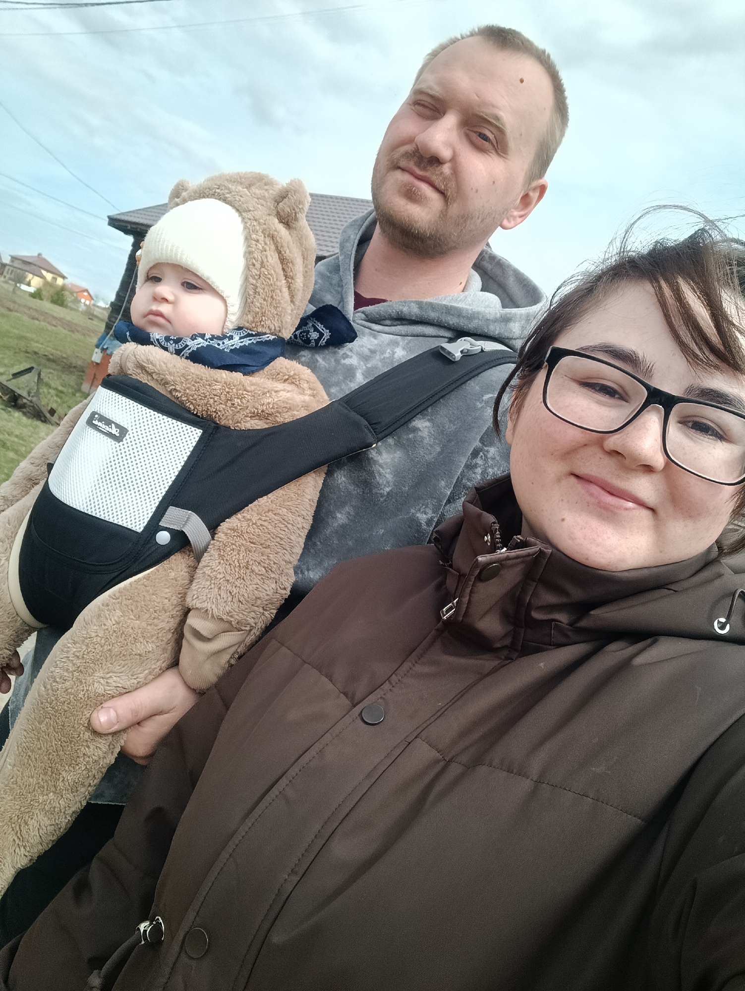 A family of three takes an outdoor selfie in Barda, Russia, under an overcast sky. In the foreground, a woman wearing black-framed glasses and a dark brown padded winter coat holds the camera, smiling faintly. Behind her, a man with light hair and a beard wears a grey hooded sweatshirt, displaying a neutral expression.

The man is carrying a baby in a black baby carrier with a white mesh panel. The brand name "i amile" is partially visible on the carrier's mesh section. The baby is dressed warmly in a light brown, fluffy bear-themed full-body suit, a white knit hat, and a blue bandana, looking contemplative.

The background shows a residential or semi-rural setting with a house featuring a dark roof and green grassy terrain with sparse vegetation to the left. All individuals are dressed in cold-weather attire, suggesting cool temperatures. The lighting is soft and diffuse, characteristic of an overcast day.