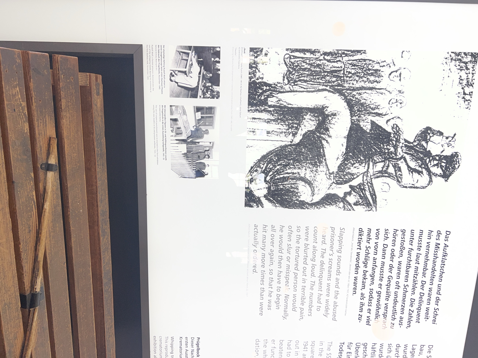 The image displays an exhibit in a museum, likely related to the Dachau concentration camp. The top portion features a black and white illustration depicting a man in a cap and uniform wielding a whip over another person who is bent over. Several other figures in uniform stand behind the victim, observing. Below the illustration, text in both German and English describes the brutality of the mistreatment, including the requirement for victims to count aloud during beatings and the potential for slurred speech due to pain.
The middle section presents two black and white photographs. The left photograph shows a person bent over a wooden structure, with other individuals in the background, suggesting a judicial or disciplinary proceeding. The caption identifies this as a former inmate, Rudolf Wolf, testifying about the use of a whipping trestle during the first Dachau trial. The right photograph depicts a recreated scene with figures in SS and inmate uniforms standing near a wooden trestle. The caption explains this is a re-enactment of corporal punishment in the first exhibition of the crematorium.
The bottom portion of the image shows a close-up of a wooden structure with horizontal slats, resembling the whipping trestle mentioned in the captions. This object is placed in the foreground, suggesting it is part of the exhibit. The overall context points to the exhibit being located at a memorial or museum in or related to Dachau, Germany, and it focuses on the corporal punishment and judicial processes associated with the former concentration camp.