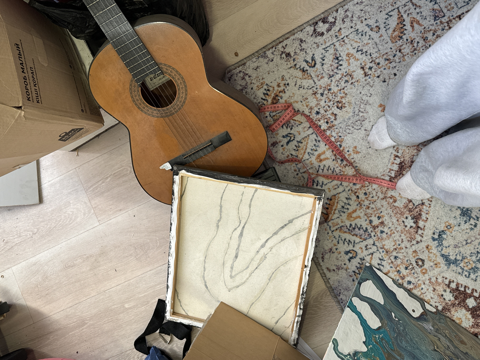 An overhead shot captures a cluttered indoor floor scene, likely in Krasnoyarsk, Russia, given the multilingual text on a box. The dominant subject is an acoustic guitar, light brown with a dark fretboard, lying on its side. Its label near the soundhole reads "alan JUANA". To the left of the guitar is a partially visible brown cardboard box, featuring text in both Russian and Kazakh: "КОРОБ МАЛЫЙ" (Small Box), "ИЗГОТОВЛЕН ИЗ 100% ПЕРЕРАБОТАННОЙ БУМАГИ" (Made from 100% Recycled Paper), "КІШІ ҚОРАП" (Small Box), and "100% КАЙТА ӨНДЕЛГЕН ҚАҒАЗДАН ЖАСАЛГАН" (Made from 100% Recycled Paper), along with a "3.5 KG" weight marking.
To the right, a patterned area rug with earthy tones, including beige, brown, blue, and orange accents, covers part of the light wooden laminate flooring. A pink measuring tape with clear numerical markings (e.g., 100, 110, 120, 130, 140, 150, 200) is sprawled across the rug. A person's lower legs and feet, clad in white socks and light grey sweatpants, are visible standing on the rug near the measuring tape.
Two artworks are also present: one canvas is overturned, revealing its wooden frame and a muted abstract design in grey lines on a light background. Another canvas is partially visible in the bottom right, displaying a vibrant abstract pour painting with blue, green, and white swirls. The scene suggests a domestic space, possibly a room where items are stored or being organized. The time of day appears to be daytime, judging by the lighting, but no specific weather conditions are discernible.