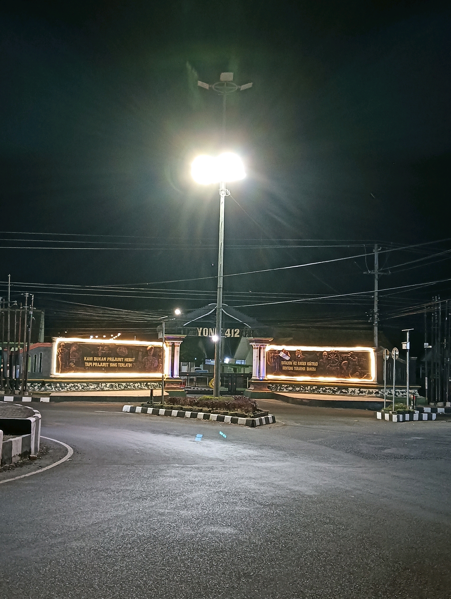 The image shows a nighttime view of the gate to Yonif 412 Raider Kostrad, located in Purworejo, Indonesia. The gate features a large, illuminated monument with bas-relief carvings depicting soldiers and military motifs. Text on the monument reads "KAMI BUKAN PRAJURIT HEBAT TAPI PRAJURIT YANG TERLATIH" on the left side and "Batalyon 412 Raider Kostrad BENTENG TERAKHIR BANGSA" on the right. The monument is lit by warm-toned lights, creating a dramatic effect against the dark sky. A tall light pole with bright floodlights stands in front of the gate, illuminating the surrounding area, including a paved road in the foreground. The road curves to the left and is bordered by a curb with black and white striped markings. Utility poles and overhead wires are visible in the background, along with the silhouette of buildings beyond the gate. The scene appears to be an entrance to a military base. There are no people visible in the image.