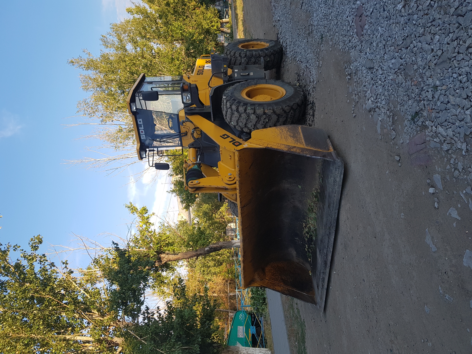 In Kurchatov, Kazakhstan, a yellow SDLG wheel loader with a large bucket is parked on a gravel and dirt surface. The loader is positioned in the foreground, angled slightly to the right. Its bucket is partially filled with green plant debris. The machine has large, rugged tires, some of which are caked with mud. The cab of the loader is visible, with windows reflecting the surroundings. Behind the loader, trees with green and autumn-colored leaves line the background. A paved walkway is visible to the left, with a portion of a green, multi-wheeled vehicle with "VÖGELE" branding on it. The sky above is clear blue with a few wispy clouds. The scene appears to be an outdoor industrial or maintenance area.