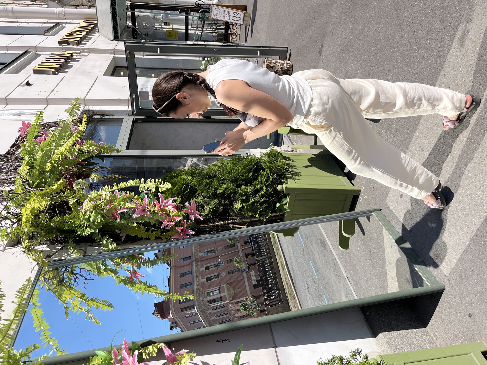 A woman with braided dark hair, wearing a white sleeveless top and cream-colored linen pants, stands on a sidewalk and looks at her mobile phone. She is holding the phone in her hands. In front of her is a large, free-standing mirror, positioned at an angle to reflect the opposite side of the street and a building. The mirror's frame is dark green, and it is adorned with hanging green ferns and pink artificial flowers.
Reflected in the mirror is a wide street with a pedestrian walkway on the left and a road with blue markings on the right. The reflected building is a multi-story structure with a distinctive pink facade, featuring ornate architectural details and rounded windows. A domed structure is visible on its roof. A decorative black metal fence runs along the sidewalk in front of the building in the reflection.
To the right of the woman and the mirror, a white building with signage in Russian (likely a shop or cafe) is visible, with its entrance doors open. A wooden sign with Cyrillic text and the number "139" is placed on the sidewalk near the entrance. Green planters containing greenery and a small conifer tree are placed on the sidewalk. The scene takes place on a sunny day, casting distinct shadows on the pavement. The location is the city of Saint Petersburg, Russia, specifically along a canal or embankment given the reflection of the building and walkway.