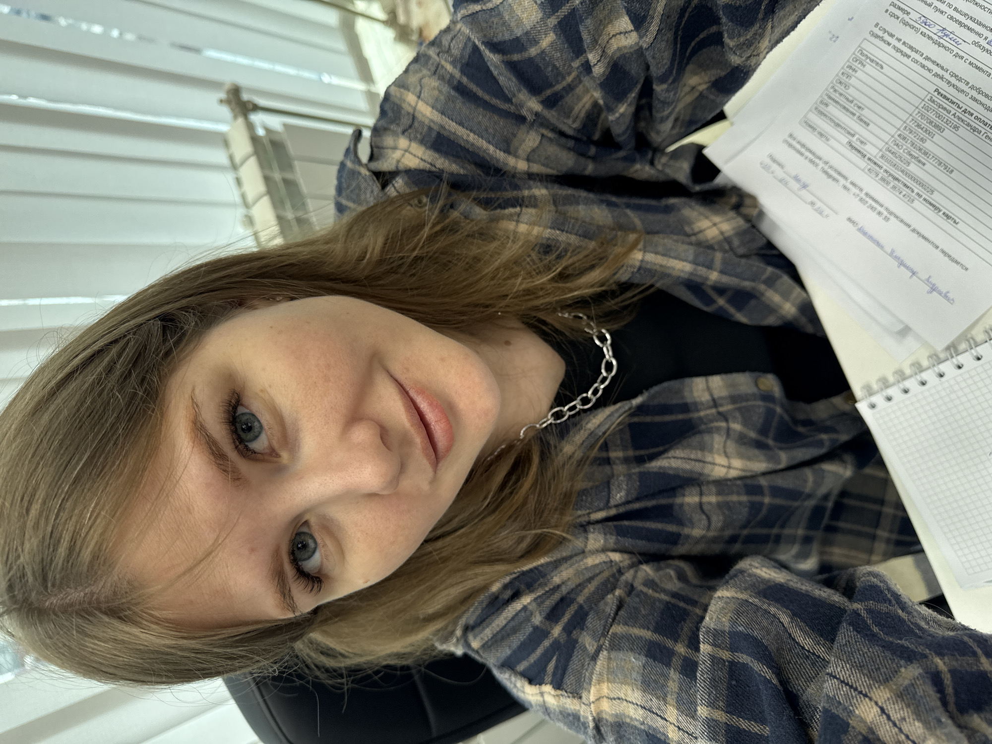 A slightly rotated, eye-level indoor shot features a young woman with light brown hair, blue eyes, and light freckles. She looks directly at the camera with a calm, neutral to slightly smiling expression. She is wearing a dark blue and tan plaid flannel shirt over a black top, accessorized with a silver chain necklace.
The setting appears to be an office or study area in Perm, Russia. In the background, a window with white horizontal blinds allows natural light into the space. To her right, a desk holds a white spiral-bound document with extensive Russian text, as well as a spiral-bound notebook.
The visible text on the document includes the heading "Реквизиты для оплаты" (Payment details). It specifies a "Получатель" (Recipient) as "Засорина Александра Гена" and lists various banking details such as TIN, KPP, OGRN, settlement account numbers, and the bank name "ПАО Сбербанк" (Sberbank). A note indicates that "Перевод можно осуществить по номеру карты" (Transfer can be made by card number). Other sections of the document discuss conditions for returning funds and legal procedures. A signature, partially visible as "Владимир", is also present. The scene suggests the woman is engaged in administrative work or reviewing financial information.