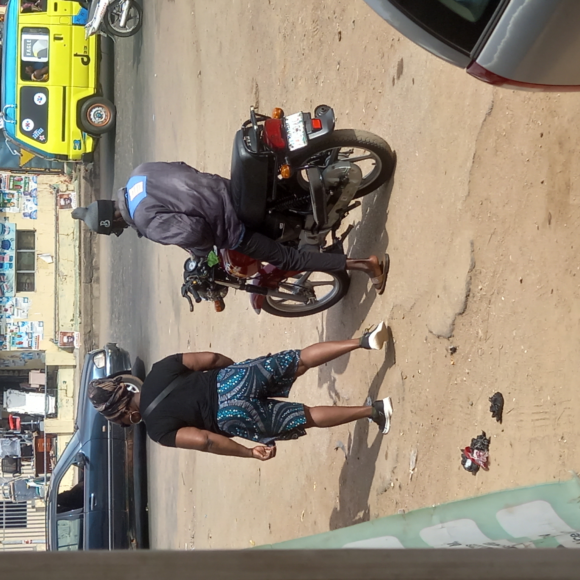 The image captures a sunlit street scene in Sardauna, Nigeria, depicting everyday activity. The ground is unpaved and dusty, with scattered debris including crumpled plastic bags.
In the foreground, a person stands on the right, facing slightly left. They wear a dark headwrap, large gold hoop earrings, a black short-sleeved top, and patterned blue and white shorts, with black and white sneakers. Their shadow is prominent on the ground behind them.
To the left, a man is seated on a dark-colored motorcycle, which has red accents. He wears a dark grey beanie, a dark jacket with a blue patch on the back, dark pants, and light-colored sandals. The motorcycle's rear license plate is mostly unreadable, but shows a taillight and amber turn signals. He appears to be stationary, possibly waiting or observing.
Various vehicles populate the background and periphery. A bright yellow commercial passenger vehicle, possibly a minibus or keke, with "EAGLE" written on its side, is visible on the far left. Another dark-colored sedan is parked on the left side of the street, and the front corner of a silver car is partially visible in the top right.
Behind the vehicles, a building facade is covered with numerous posters, some displaying faces, likely for political campaigns or local events. The scene suggests a busy, urban or semi-urban environment under clear, sunny skies, indicated by the harsh shadows. A green strip at the bottom of the image partially reveals the letters "ICE".
