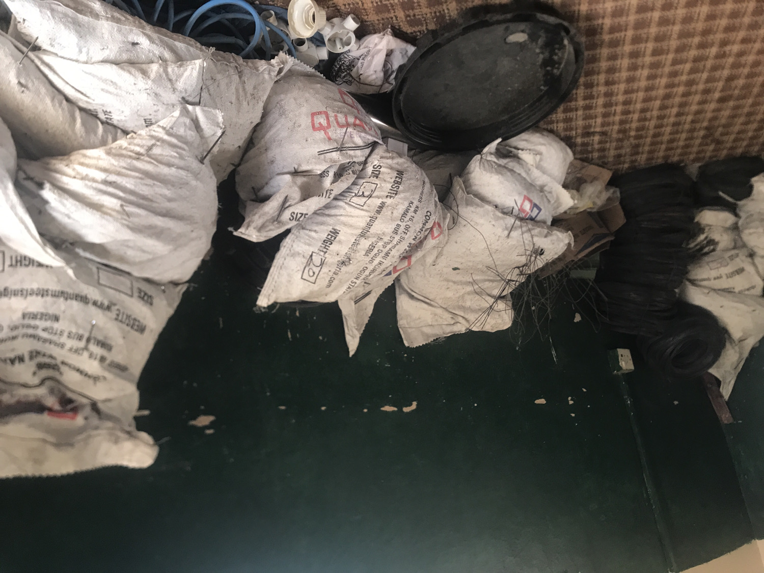 The image captures a cluttered indoor scene, primarily filled with numerous white sacks or bags piled together. These sacks appear to be made of woven plastic and are somewhat dirty and creased, suggesting they contain materials or goods. Many of them display printed text and logos.
Among the sacks, various other items are visible. Towards the upper left, blue coiled hoses or pipes are intertwined with some of the bags. In the upper right, a black, round object resembling a lid or shallow pan rests on a brown checkered surface, which looks like a mat or carpet. Adjacent to this, more white plastic pipe fittings are scattered. On the right side of the image, below the black lid, several coils of black material, possibly electrical wires or rubber hoses, are neatly stacked or bundled. A dark green floor occupies the lower and central part of the frame, showing some specks of dirt and wear.
No human activity is depicted, and the setting appears to be a storage room, workshop, or an area where materials are kept. The lighting is ambient, without clear indications of the time of day.
Visible text on the white sacks provides specific details:
- "WEBSITE www.quantumsteelsnigeria.com"
- "NIGERIA"
- "OGUN STATE"
- "KAMALO BUS STOP"
- "ADDRESS KM 16, OFF SHAGAMU IKOROBO"
- "SIZE" (followed by numbers like "3")
- "WEIGHT" (followed by numbers like "20")
- The word "COMMON" and possibly "QUANTUM" as part of a logo or brand name.
These details indicate the origin or association of the contents within the sacks, pointing to a location in Nigeria.